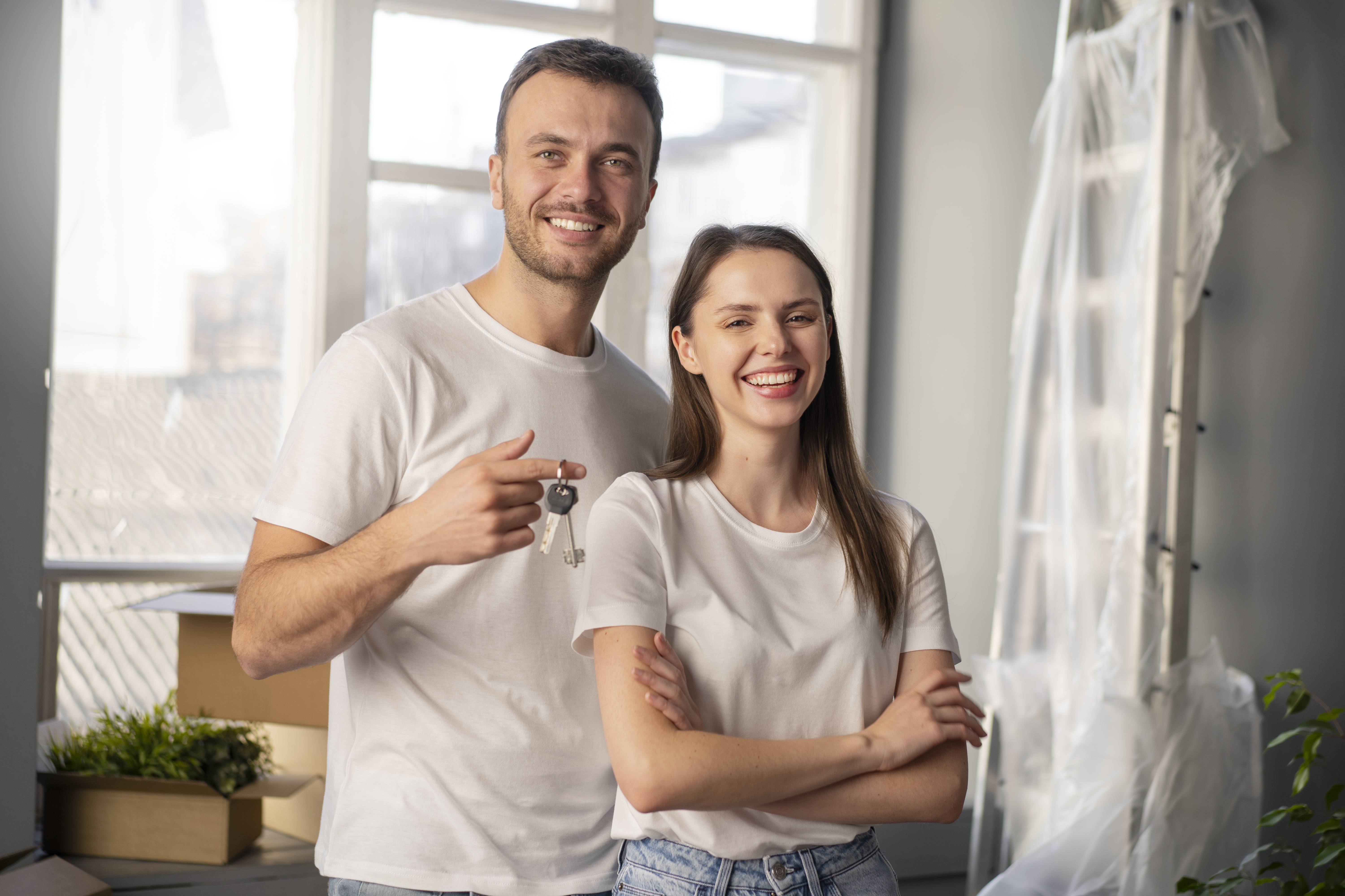 A smiling couple stands together in a bright room filled with moving boxes and a ladder; the man holds up a set of keys while the woman stands with her arms crossed, indicating they are moving into a new home.