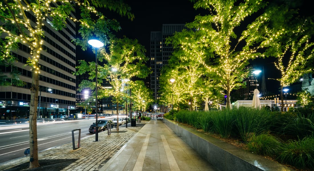 A vibrant city street illuminated by twinkling lights wrapped around rows of trees, creating a warm and inviting glow against the sleek modern buildings.