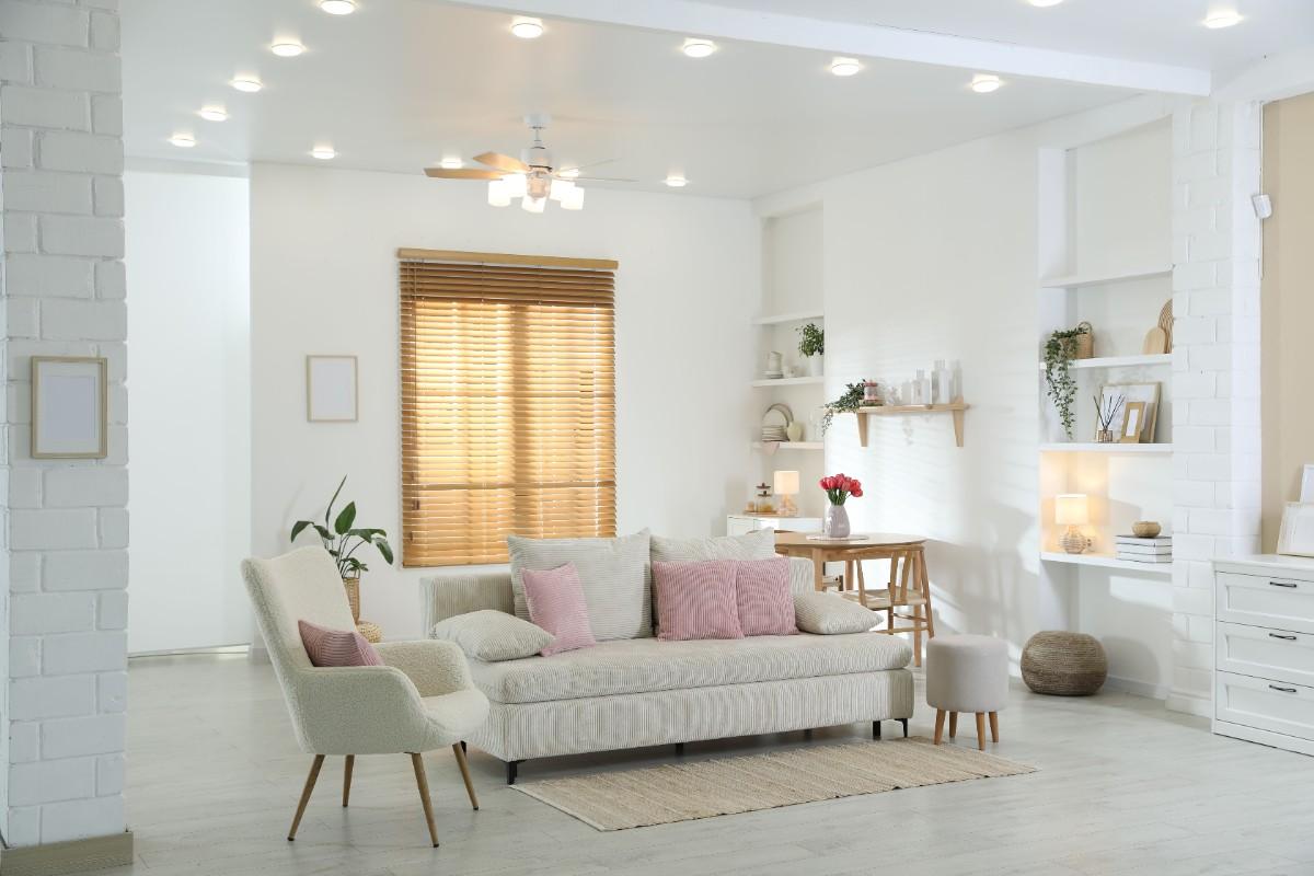 A cozy, modern living room with a white sofa, pink cushions, and a beige armchair. Warm lighting, wooden blinds, and plants create a calm and inviting atmosphere.