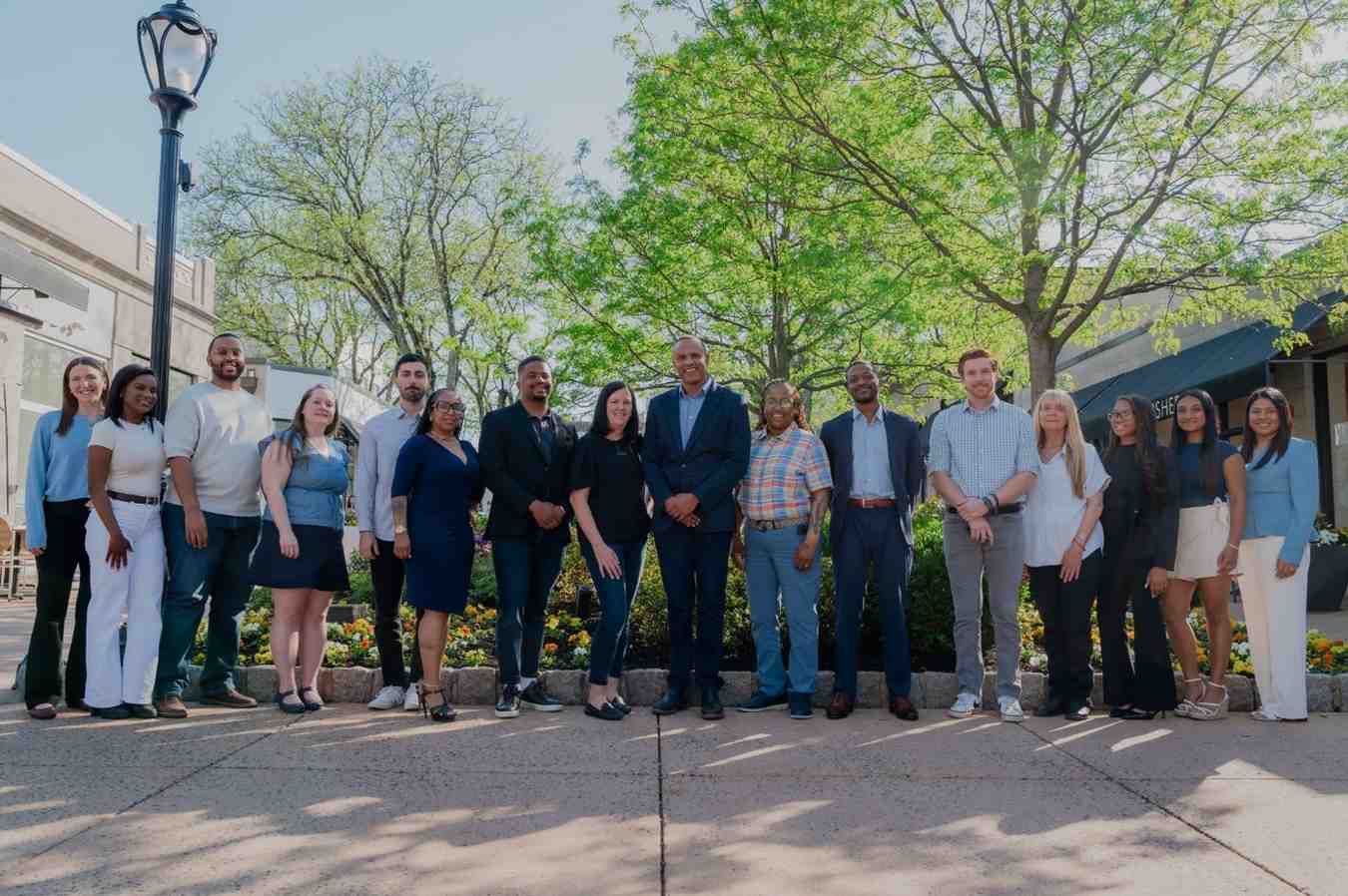 A diverse group of 16 people stands in a line outdoors on a sunny day, with trees and buildings in the background, expressing unity and positivity.