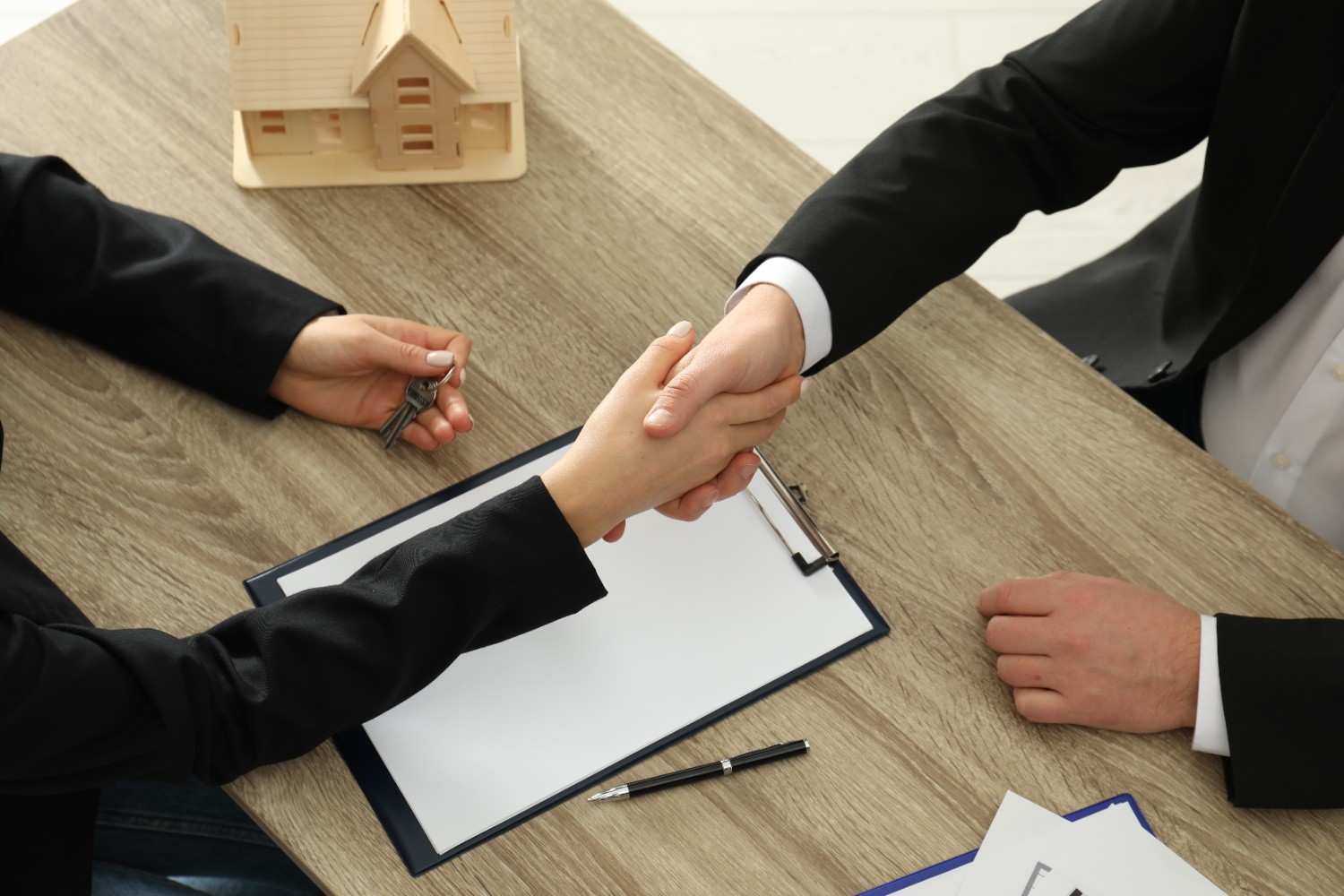 A home buyer and real estate agent shaking hands after closing, with documents and keys laid out on the desk.