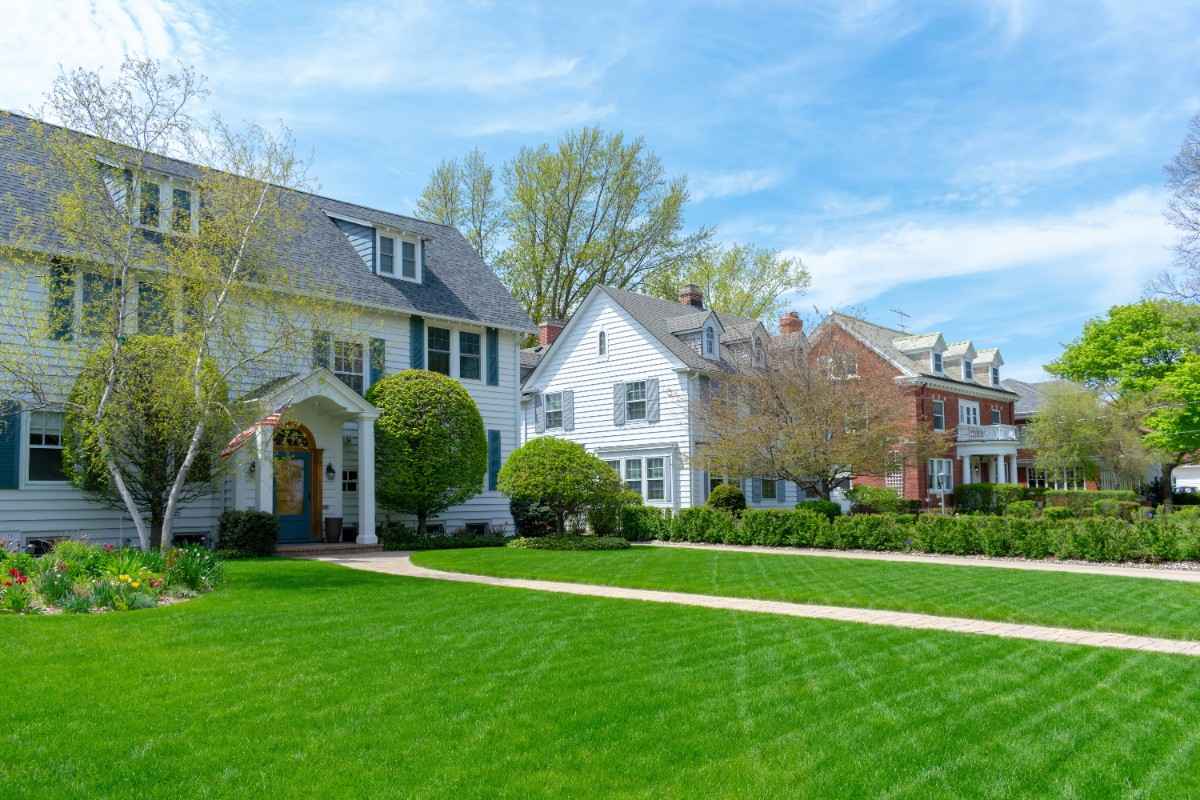 A row of elegant suburban homes with manicured lawns and trees under a bright blue sky, evoking a serene, picturesque neighborhood setting.