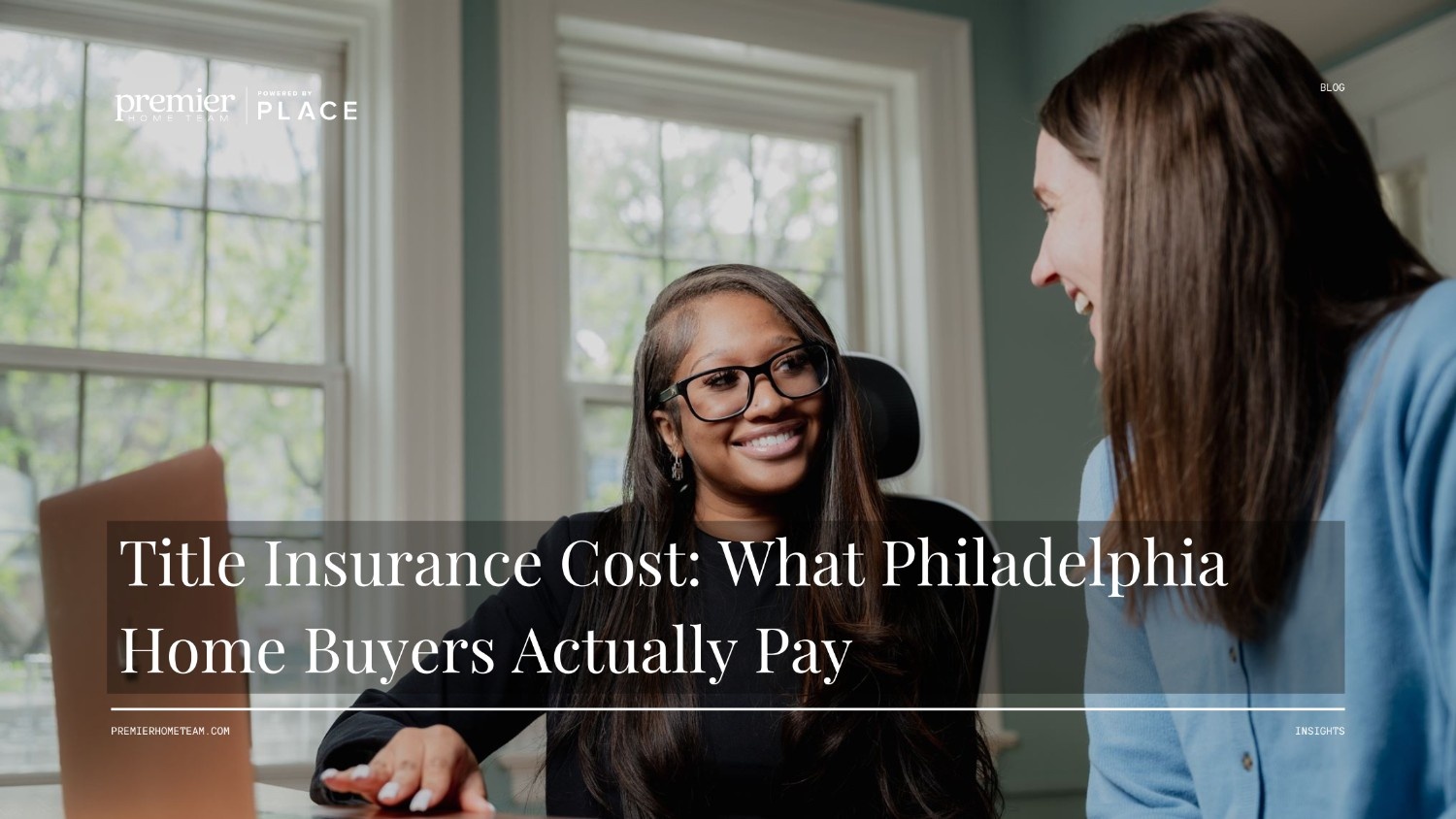 A real estate blog header image featuring a full-width photo background with a subtle dark overlay. The scene shows two women sitting together at a table in a brightly lit room. A Black woman with glasses and long dark hair sits in an office chair, smiling warmly toward the woman next to her. A white woman with long brown hair wearing a light blue sweater smiles back in profile. Bold white serif text overlaid across the bottom reads, 