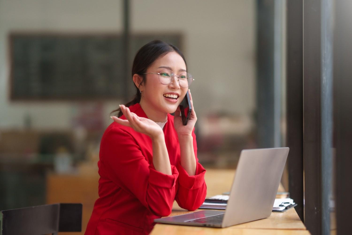 A young professional woman smiling and gesturing excitedly during a phone call while looking at her laptop. This represents a recruit's positive experience during their first discovery call with the Premier Home Team.