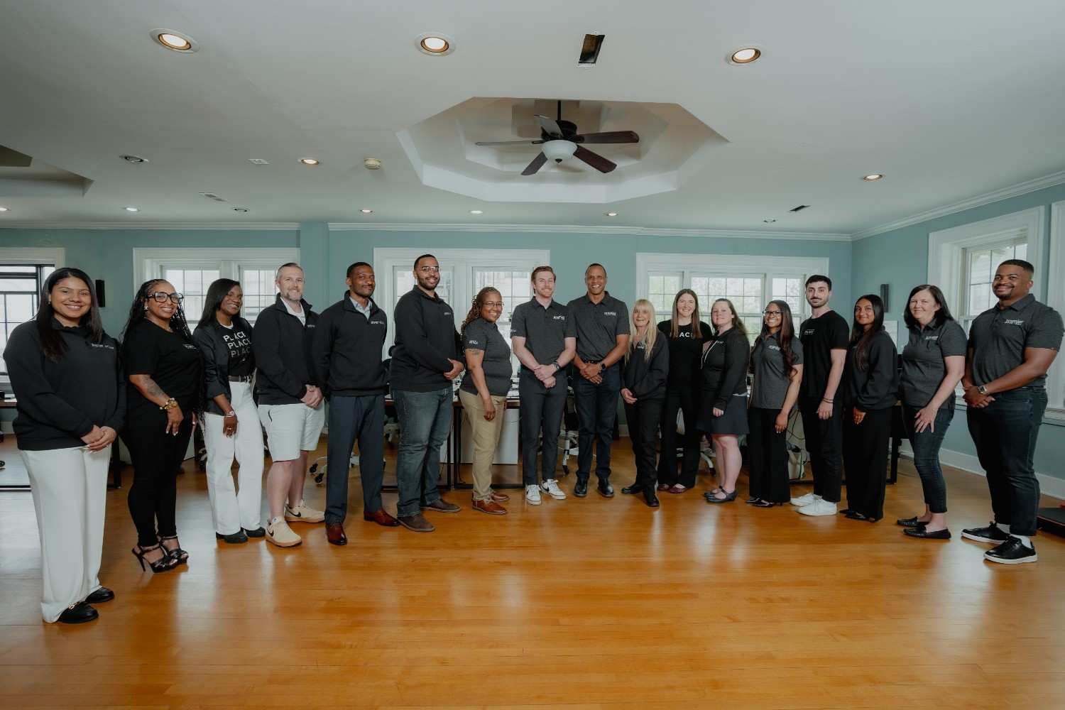 A diverse group of fifteen people stand in a spacious, well-lit room with wooden floors. They are dressed in business casual attire, smiling warmly.