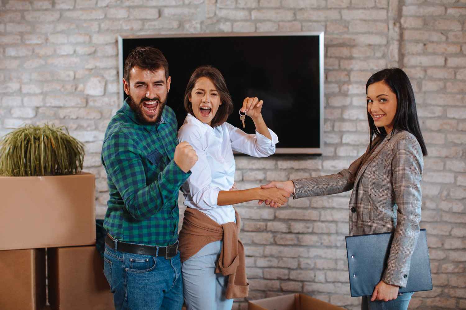 Two women shake hands in front of a TV; one holds a key, the other a folder, while a man looks ecstatic nearby.