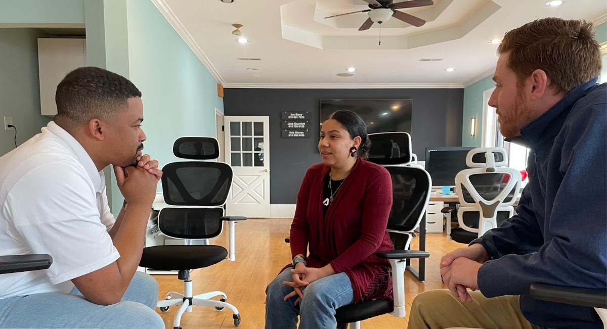 Three people sit in swivel chairs having a discussion in an office space with a relaxed, collaborative atmosphere.