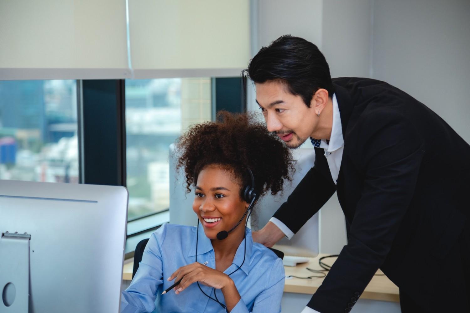 A smiling woman in a headset sits at a computer, assisted by a man leaning over.