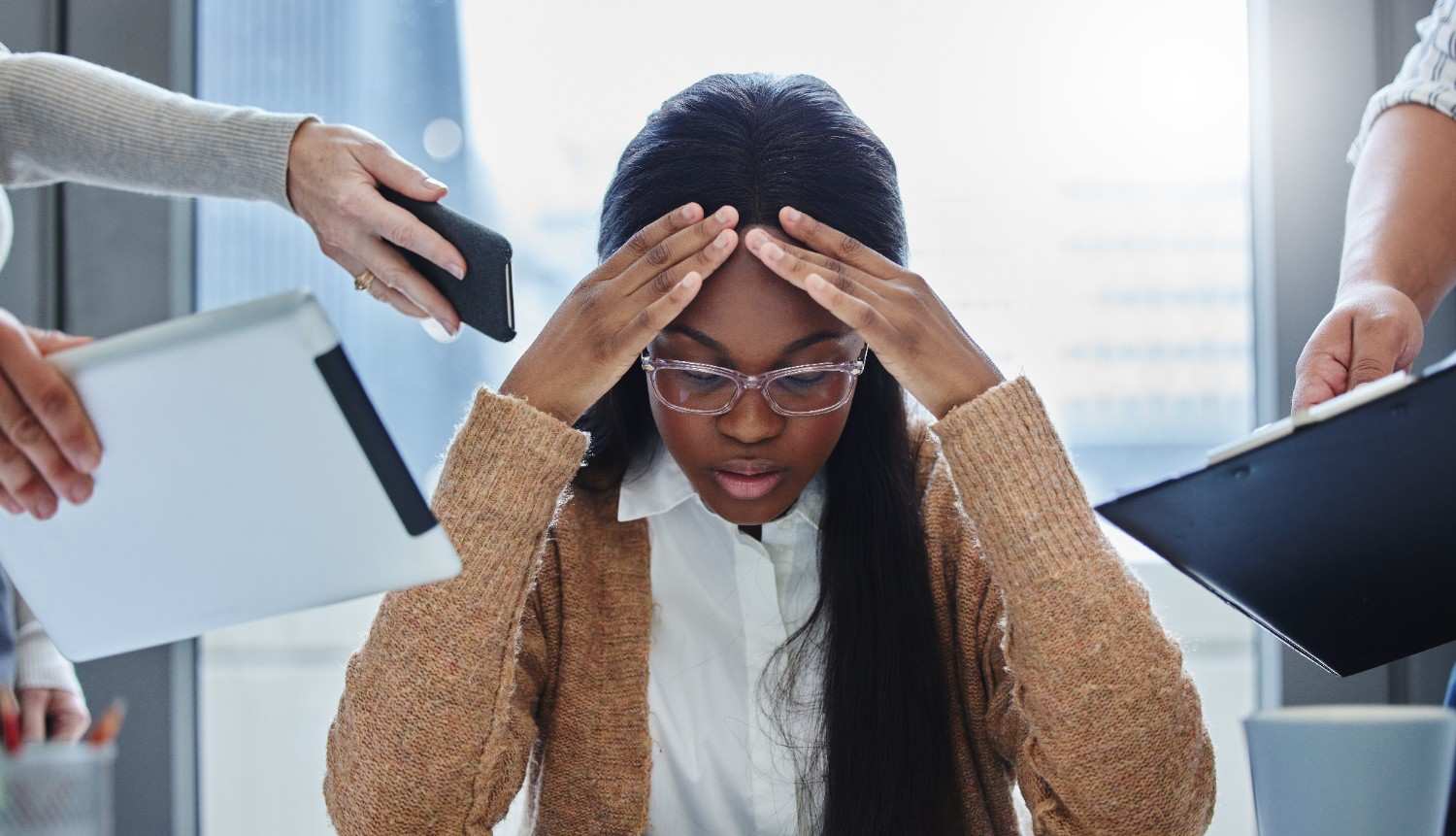 A woman sits at a desk, holding her head in her hands, looking stressed or overwhelmed.