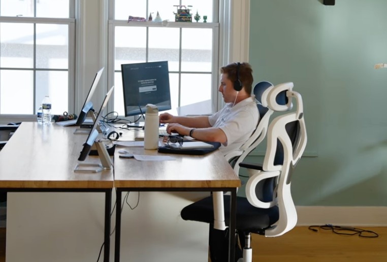 A real estate professional with headphones working on a computer at a modern wooden office desk next to bright windows.