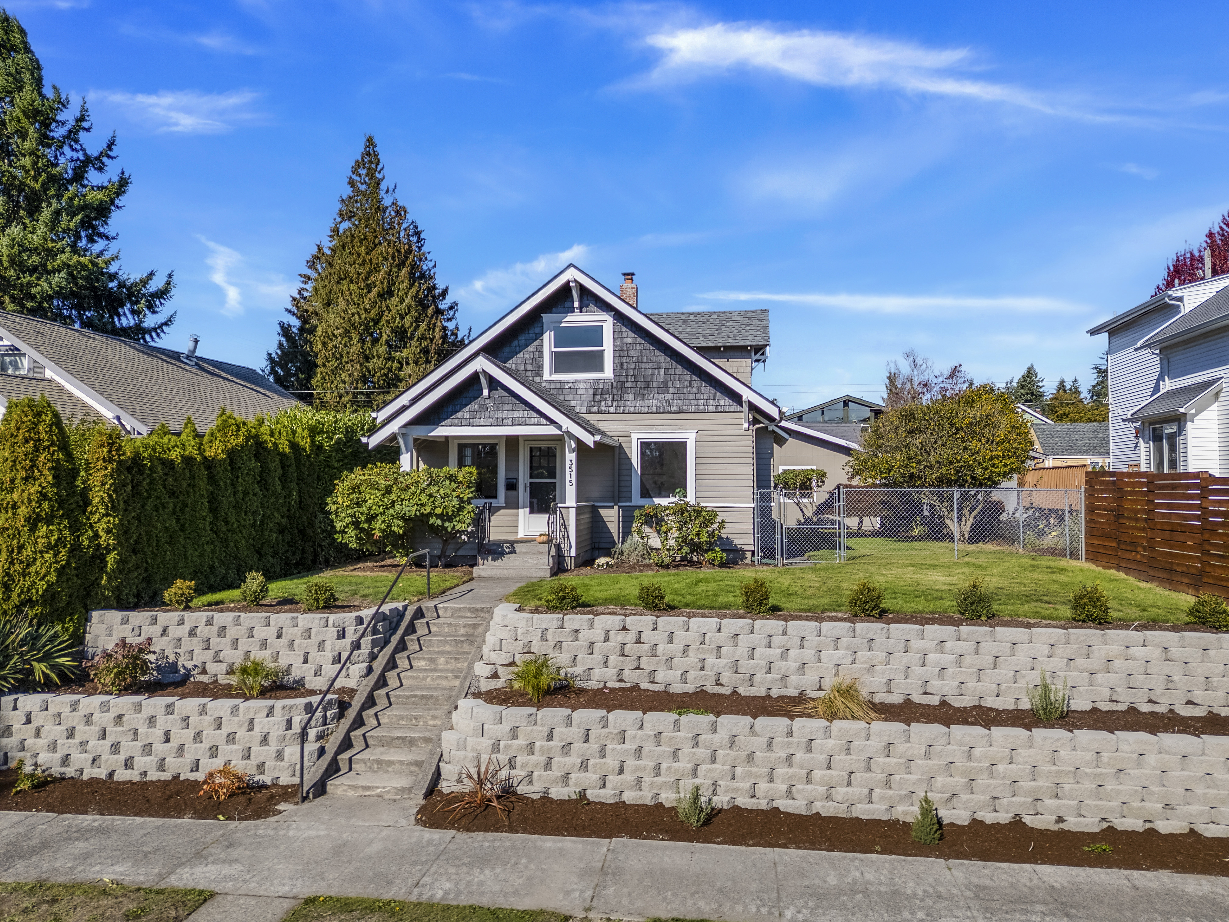Front view of a charming gray Craftsman-style home with white trim, a covered front porch, and a tiered retaining wall landscaped with shrubs and young plants. The house sits on a raised corner lot with a neatly maintained lawn, surrounded by neighboring homes and tall evergreen trees under a bright blue sky.