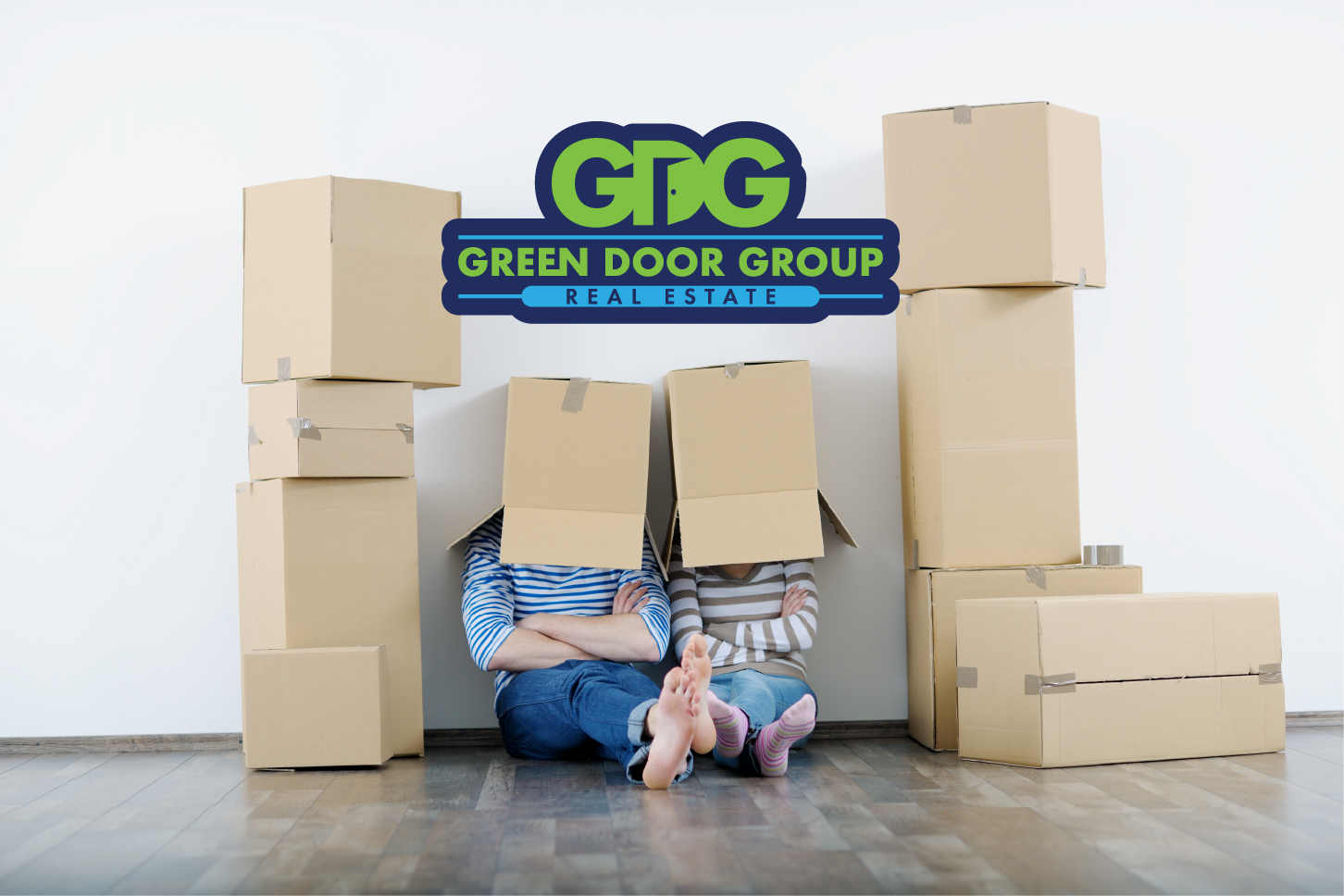 Two people with cardboard boxes over their heads sitting on a hardwood floor amid moving boxes