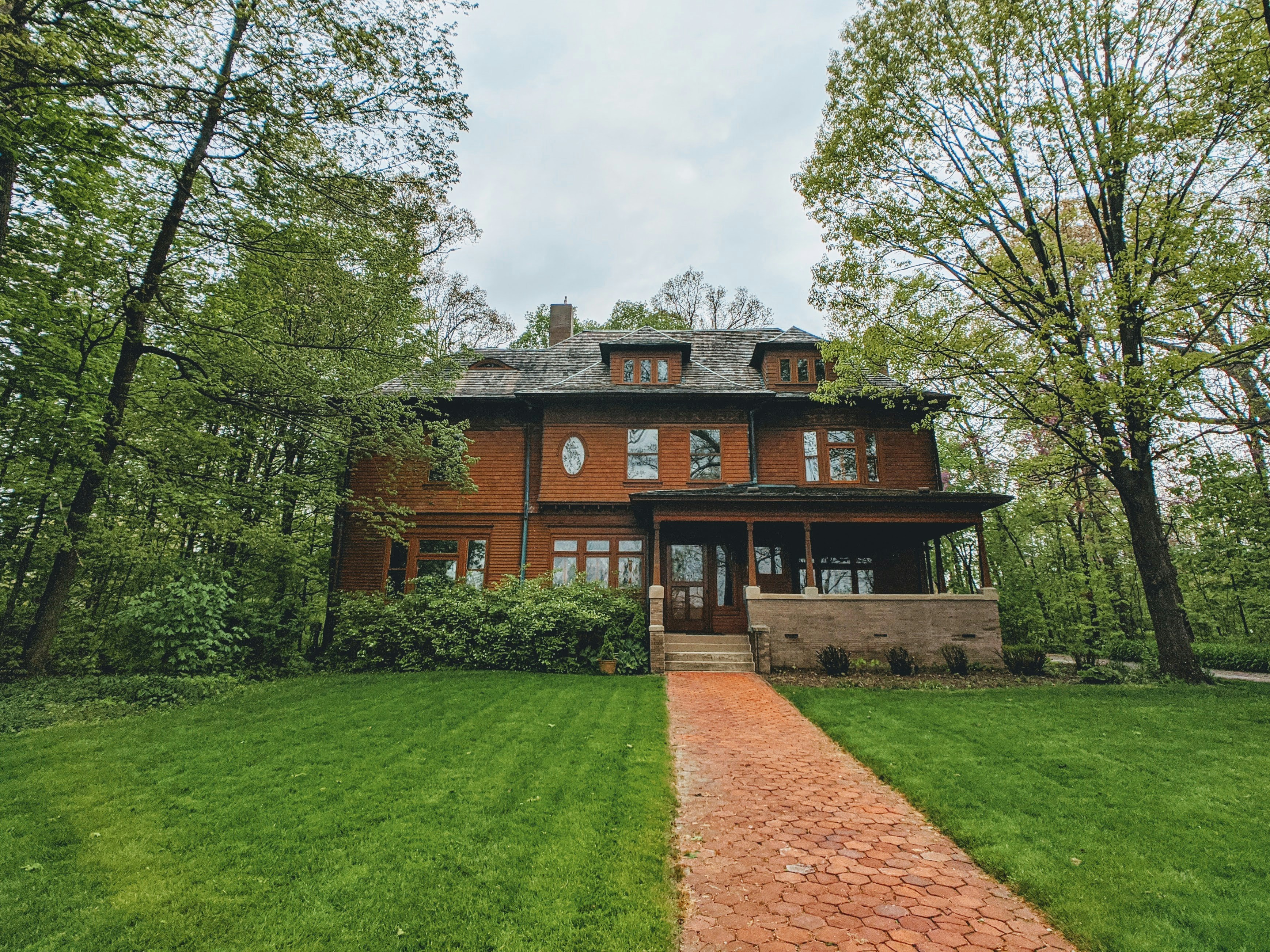 Exterior view of a large older two-story home with a front porch, surrounded by trees and a brick walkway leading to the entrance