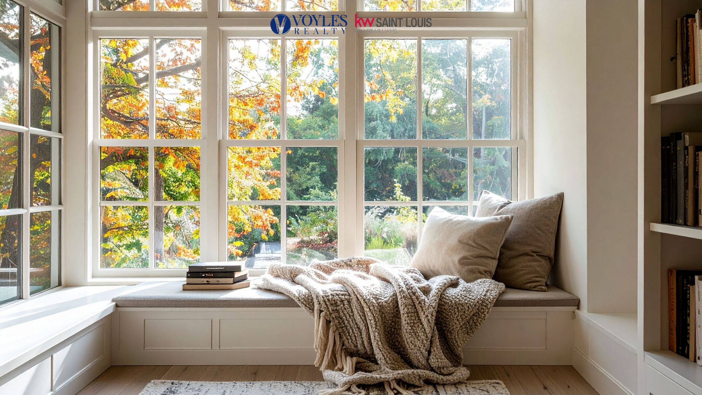Cozy indoor window seat with a knit blanket, pillows, and stacked books, looking out a large window at vibrant yellow and orange autumn trees. Voyles Realty and KW Saint Louis logos are visible at the top.