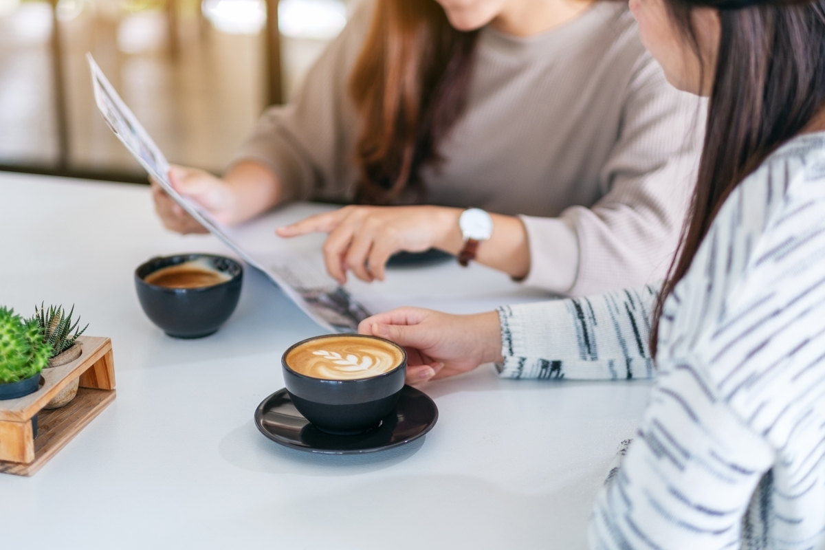 Two people sitting at a white table in a bright cafe, discussing a document over lattes. One person points to the paper while the other holds a black coffee cup with latte art, creating a collaborative and professional atmosphere.