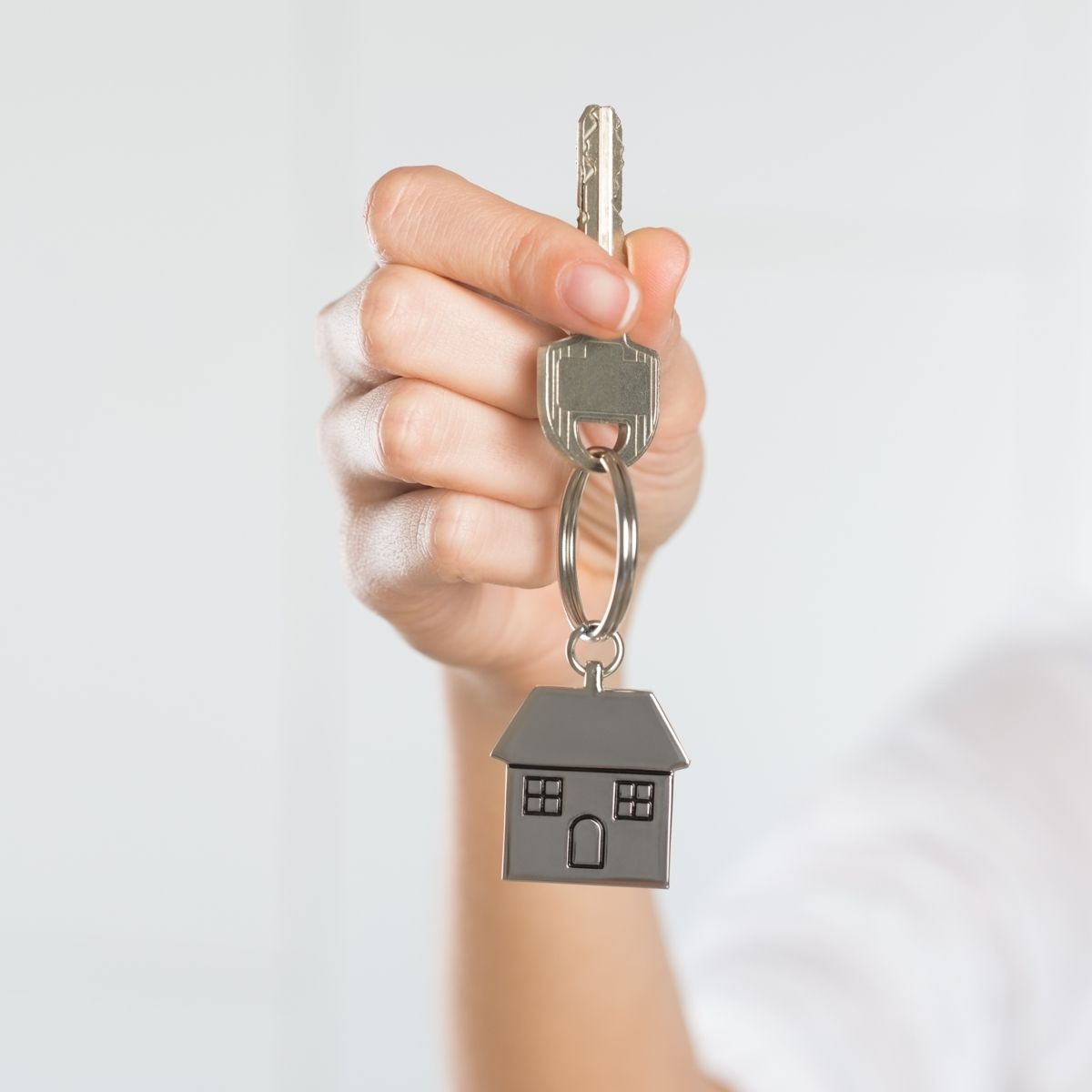 A person's hand holding up a silver house key on a keyring with a small, metallic house-shaped keychain against a blurred, plain white background.