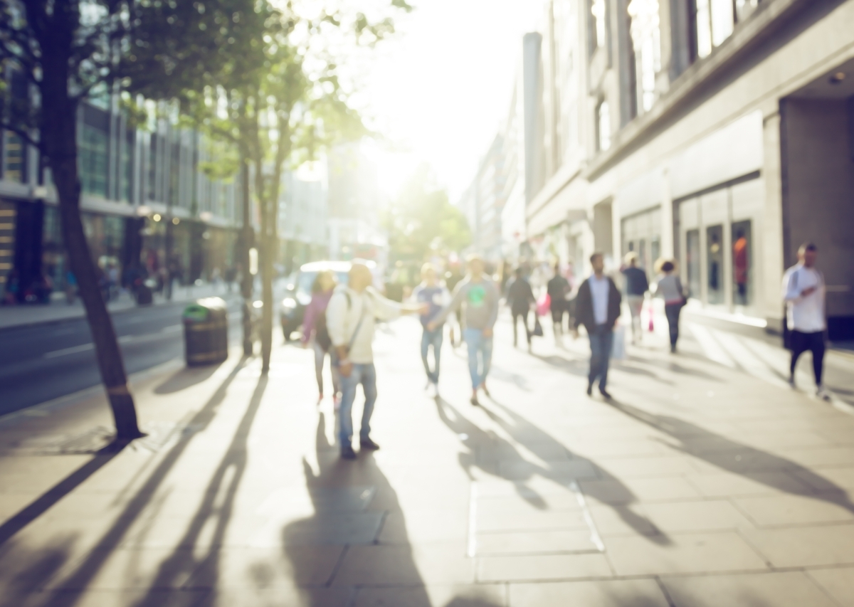 Blurred image of pedestrians walking along a sunny city sidewalk lined with trees and shops, casting long shadows.