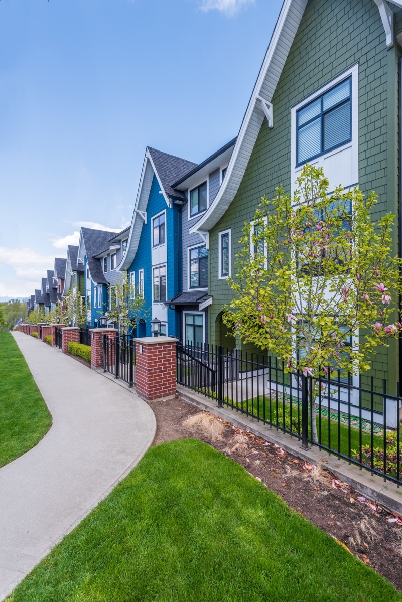 A row of colorful, modern townhouses with steep gabled roofs, featuring green and blue siding, lined by a concrete walkway and a black metal fence
