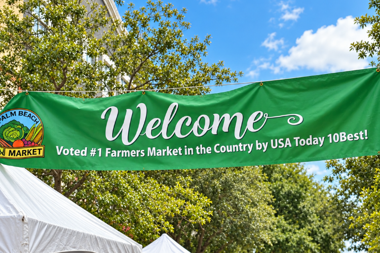 Green welcome banner at the West Palm Beach GreenMarket noting its USA Today 10Best No. 1 farmers market ranking, above white vendor tents