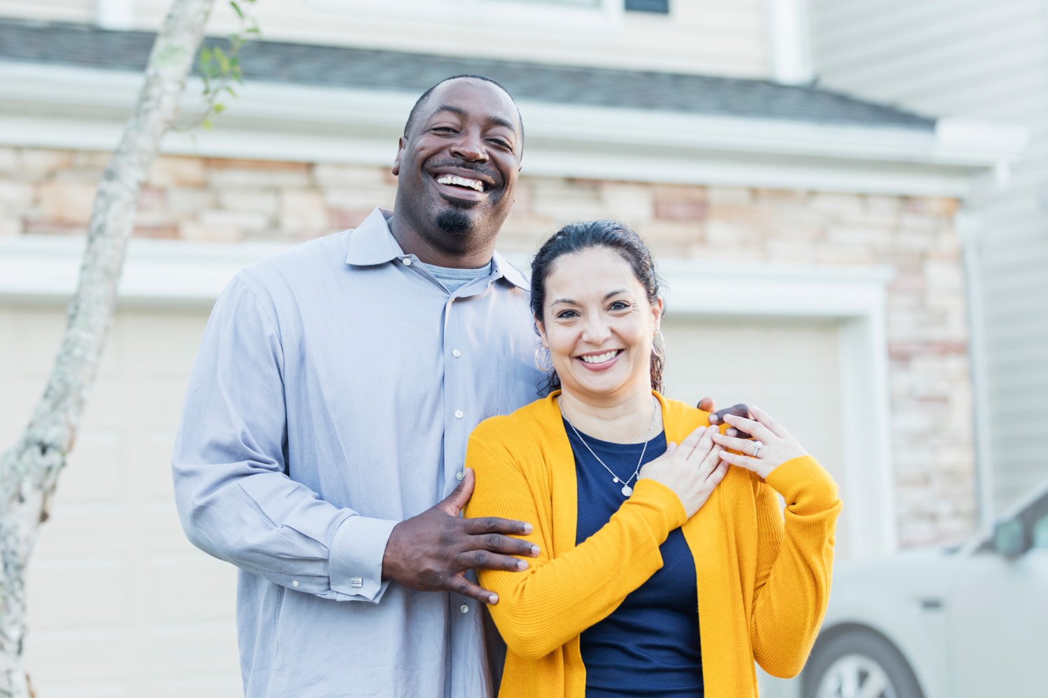 A mature interracial couple standing together outside their home, car in the driveway, smiling at the camera.