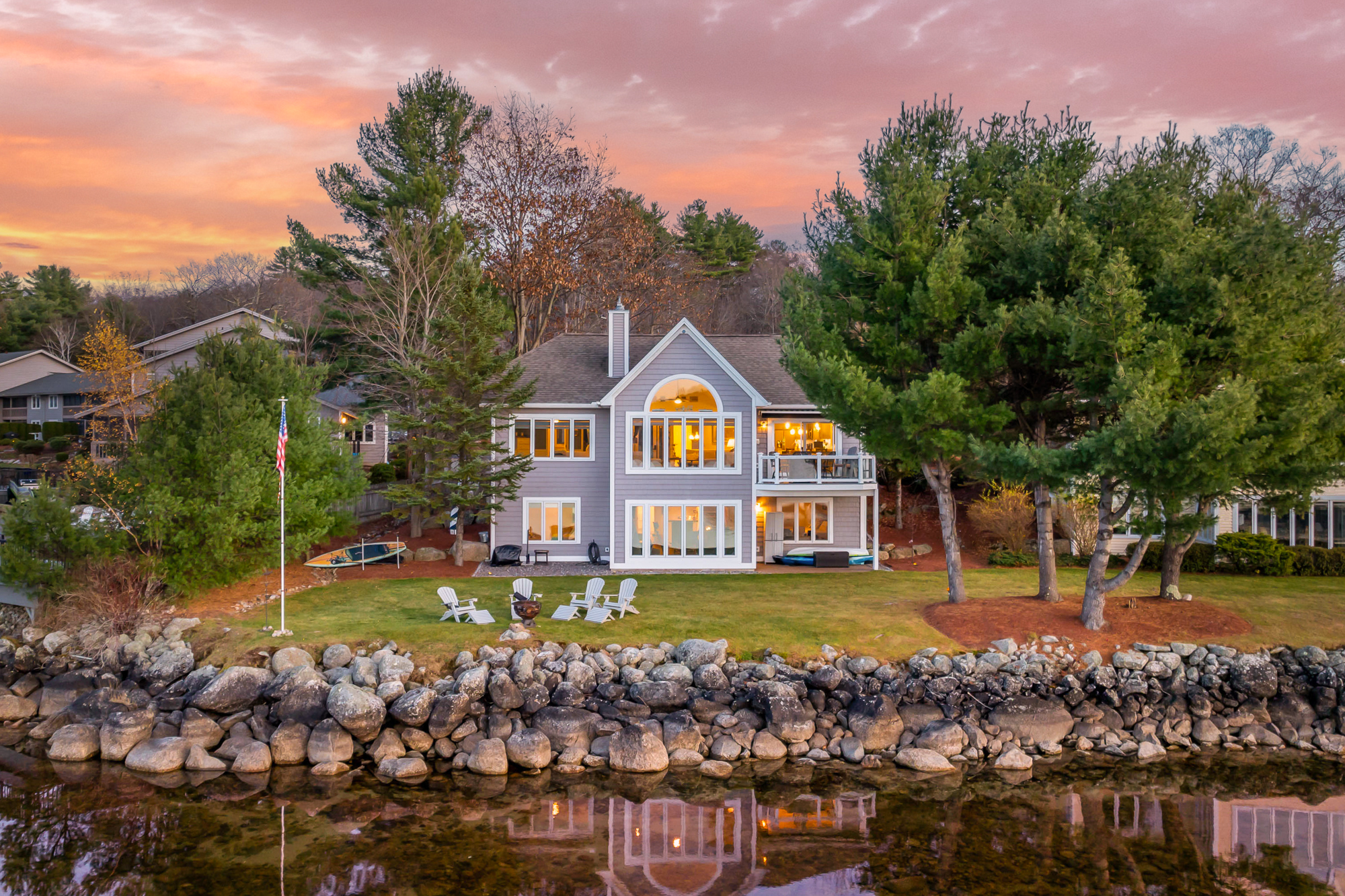 Waterfront lake house in Laconia, NH at sunset, featuring a modern two-story design with large windows, balcony, lawn, Adirondack chairs, fire pit, and stone shoreline.