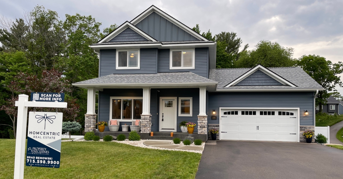 A Homcentric Real Estate for sale sign in front of a blue craftsman-style home in the Chippewa Valley, representing the Eau Claire Wisconsin housing market in 2026