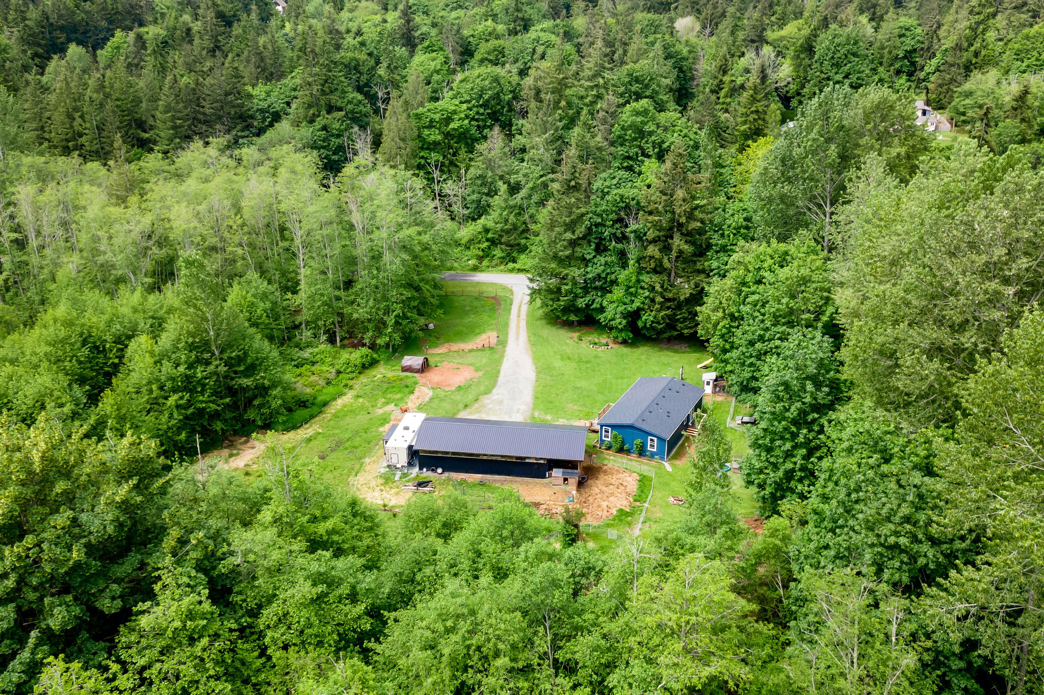 view of 1707 Bonnie Lane on Camano Island featuring a well-maintained manufactured home, spacious gravel driveway, and large detached 3-bay shop surrounded by trees and greenery.