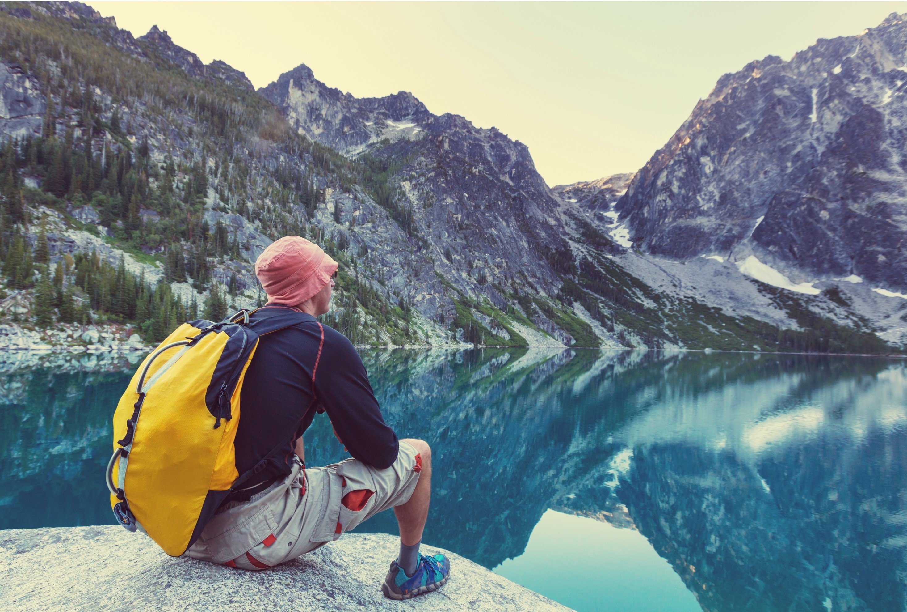 A hiker wearing a yellow backpack and pink hat sits on a rock, overlooking a serene alpine lake surrounded by steep, rugged mountains. The clear blue water reflects the dramatic peaks and evergreen trees, capturing the tranquil beauty of a summer day in the Pacific Northwest.