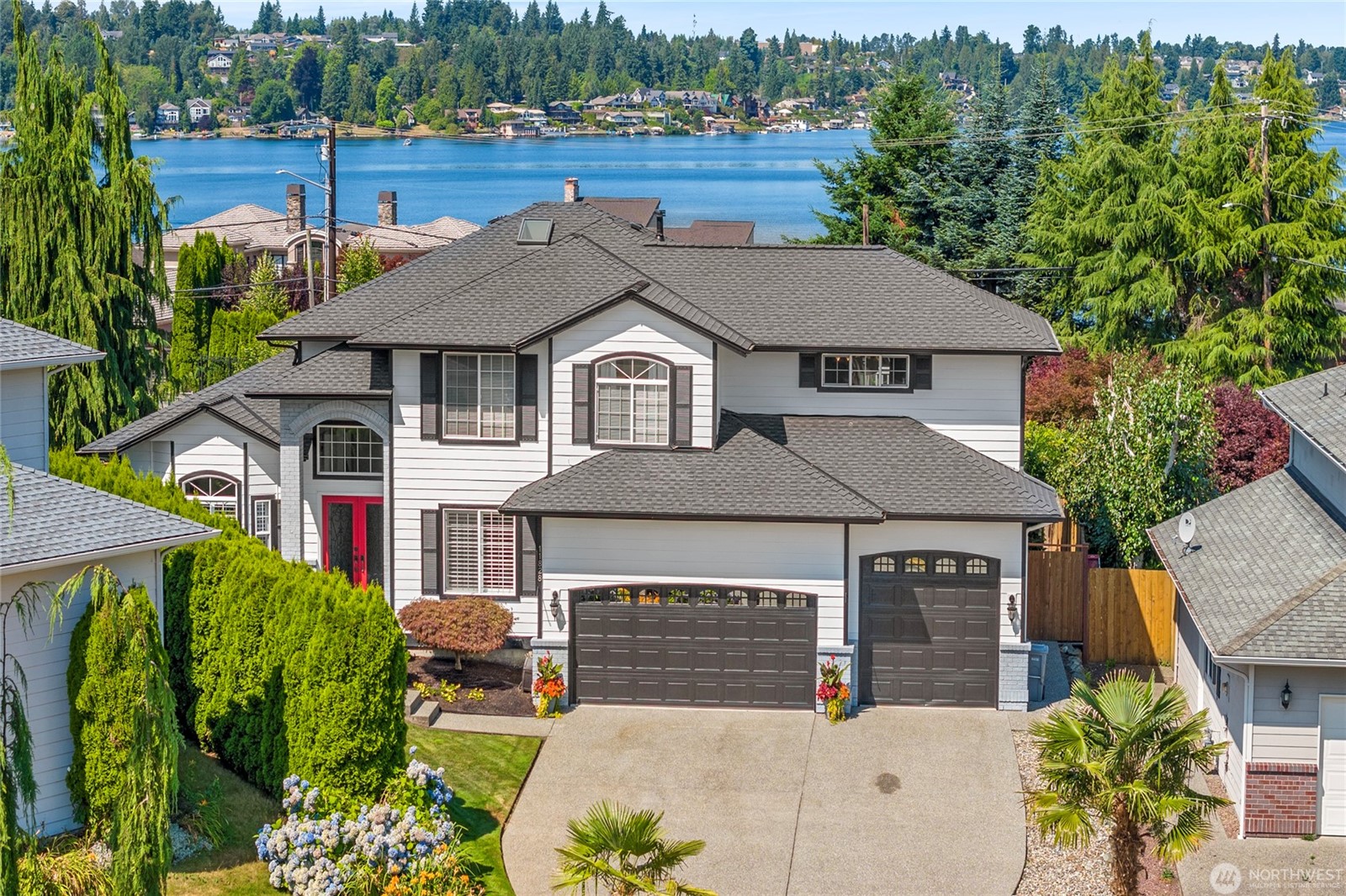 Two-story modern home with black shutters and red front door, featuring a three-car garage and well-manicured landscaping, set against a backdrop of Lake Stevens with forested hills and waterfront homes in the distance.