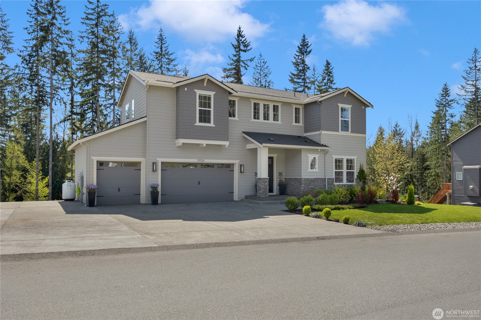 Two-story modern home with three-car garage, gray siding, and stone accents, located on a quiet residential street with a manicured front lawn and surrounded by tall evergreen trees in Lake Stevens, Washington