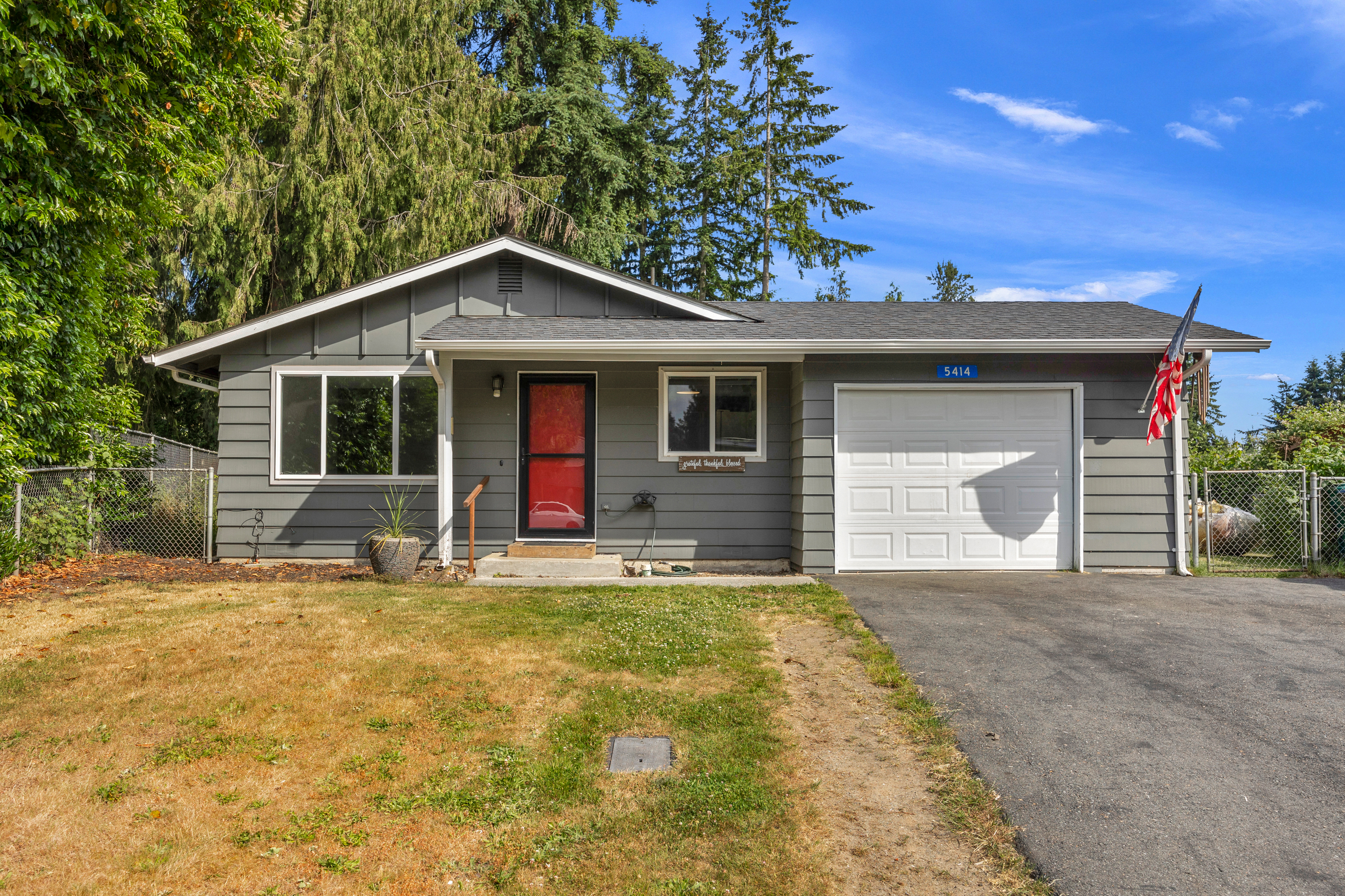 Single-story gray house with a red front door, white garage door, and American flag, set against a backdrop of tall evergreen trees on a sunny day. A paved driveway and partially fenced yard with a mix of green and dry grass complete the front exterior.