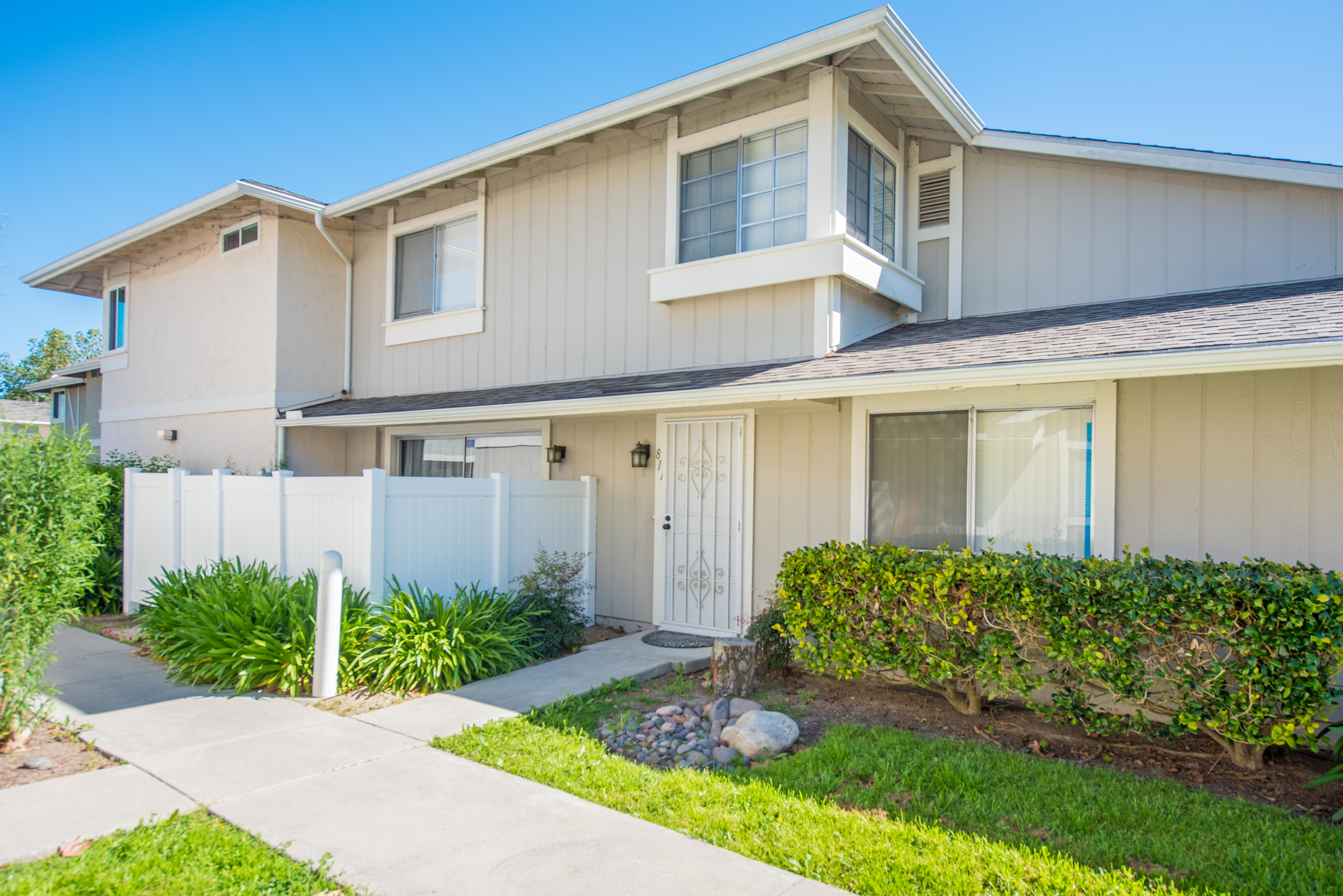 Exterior view of a two-story beige condominium at 811 Pillar Point Way in Oceanside, California featuring a private front entry with security door, white vinyl fencing, landscaped greenery, and a concrete walkway leading to the unit. 
