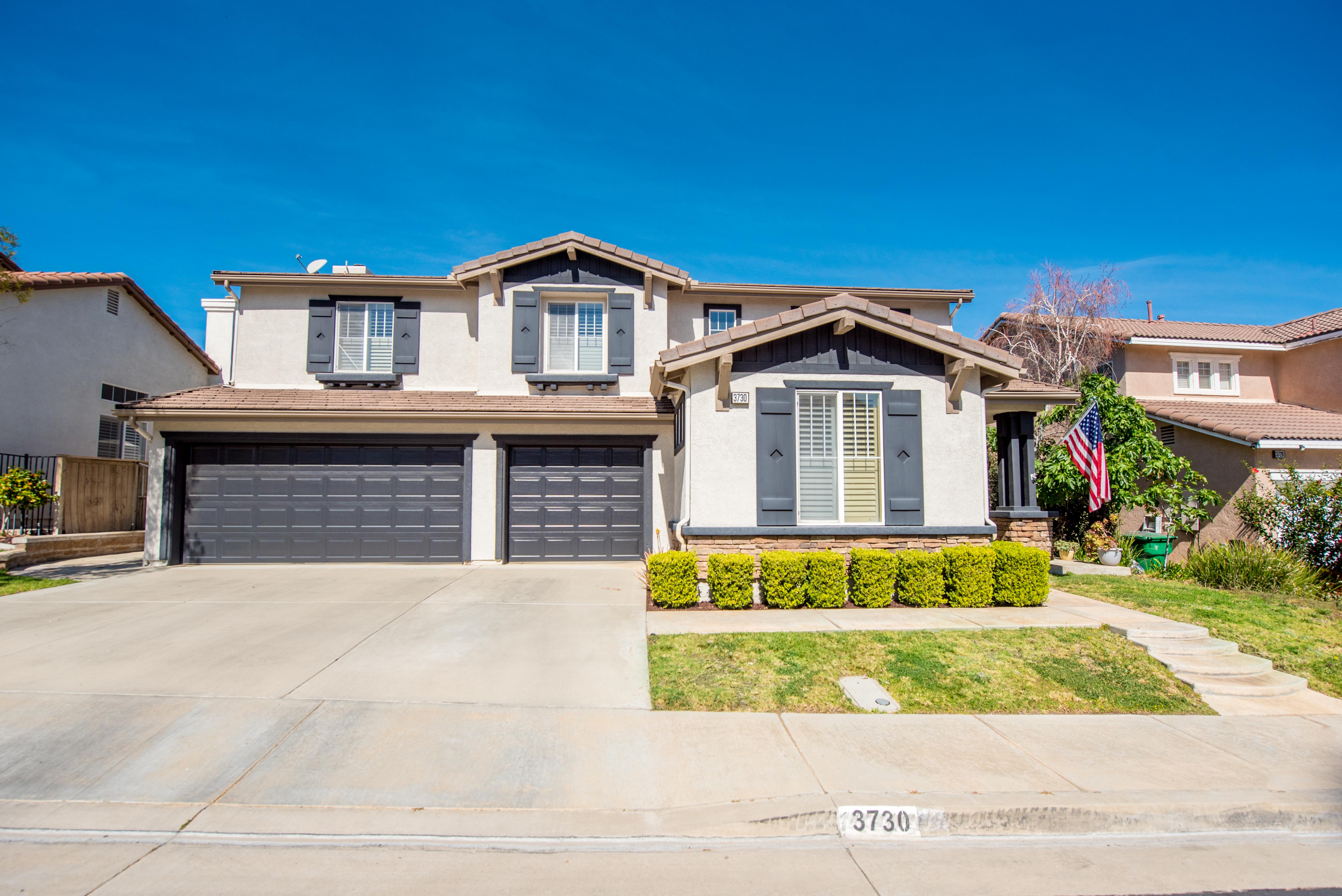 Front exterior of two-story home at 3730 Wallowa Circle in Corona, California featuring a three-car garage, light stucco exterior with dark shutters, landscaped front yard, and an American flag near the entry. 