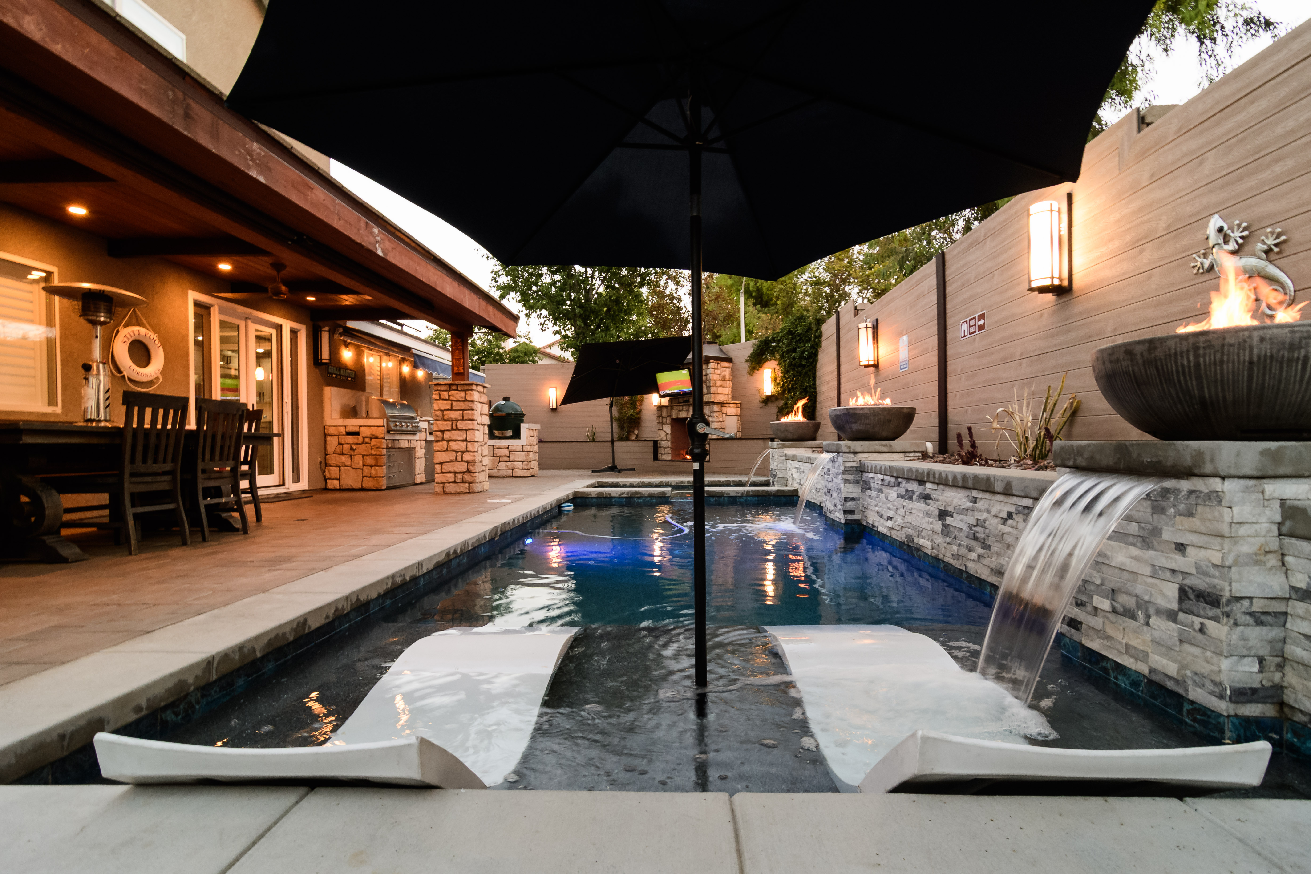Residential backyard with a swimming pool, built-in lounge chairs, water features, fire bowls, and a covered patio. 