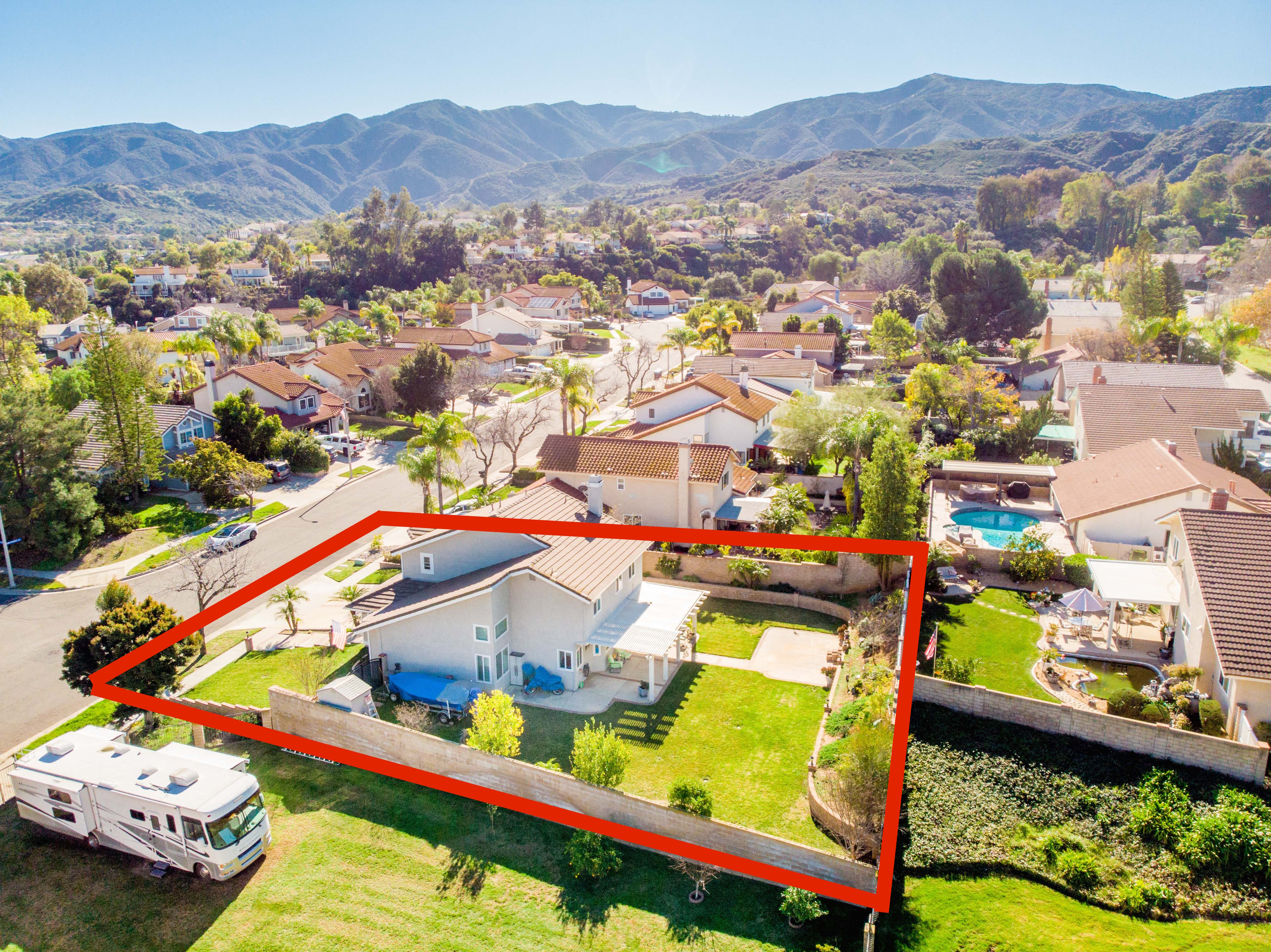 Aerial view of a residential home in Corona, California with a fenced backyard and RV Parked along side of the property. The home is outlined in red, with surrounding neighboring houses and nearby hills visible in the background. 