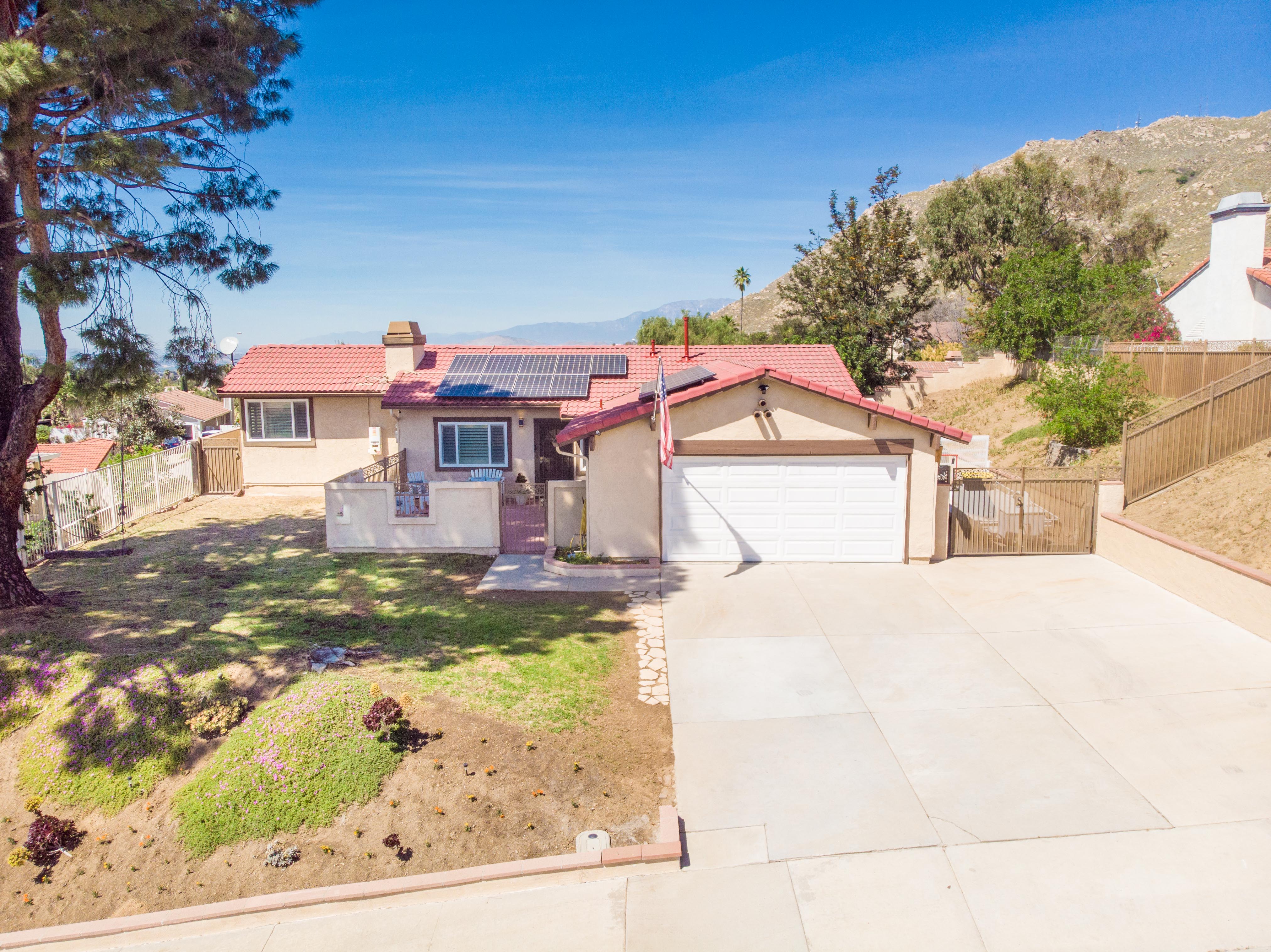Front aerial view of 11703 Black Hawk Lane in Moreno Valley featuring a single-story home with solar panels, mountain backdrop, spacious driveway, and a quiet residential setting.