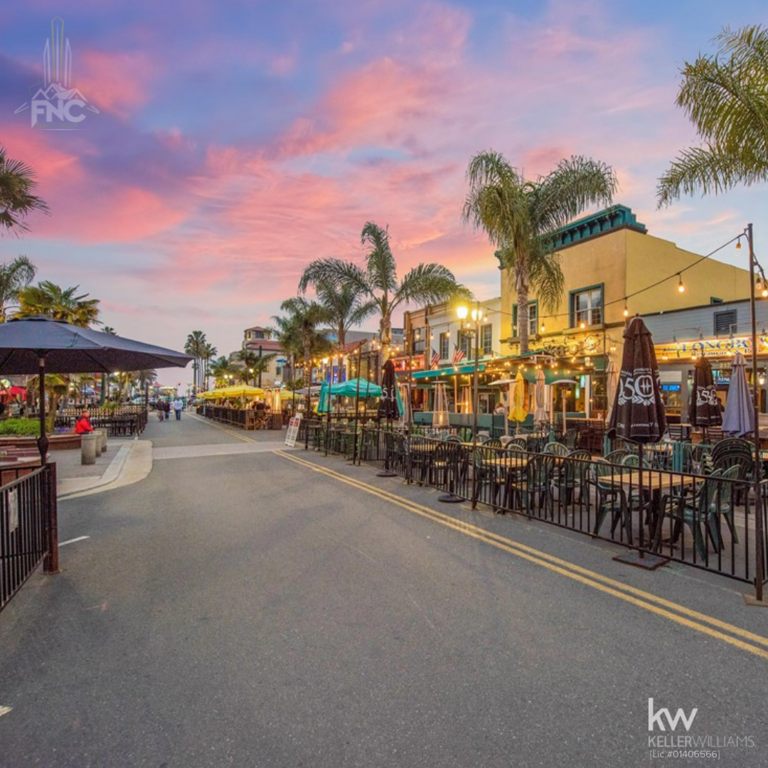 Street view of downtown Huntington Beach, California, showing outdoor restaurant seating, palm trees, storefronts with string lights, and a pedestrian friendly roadway at sunset.