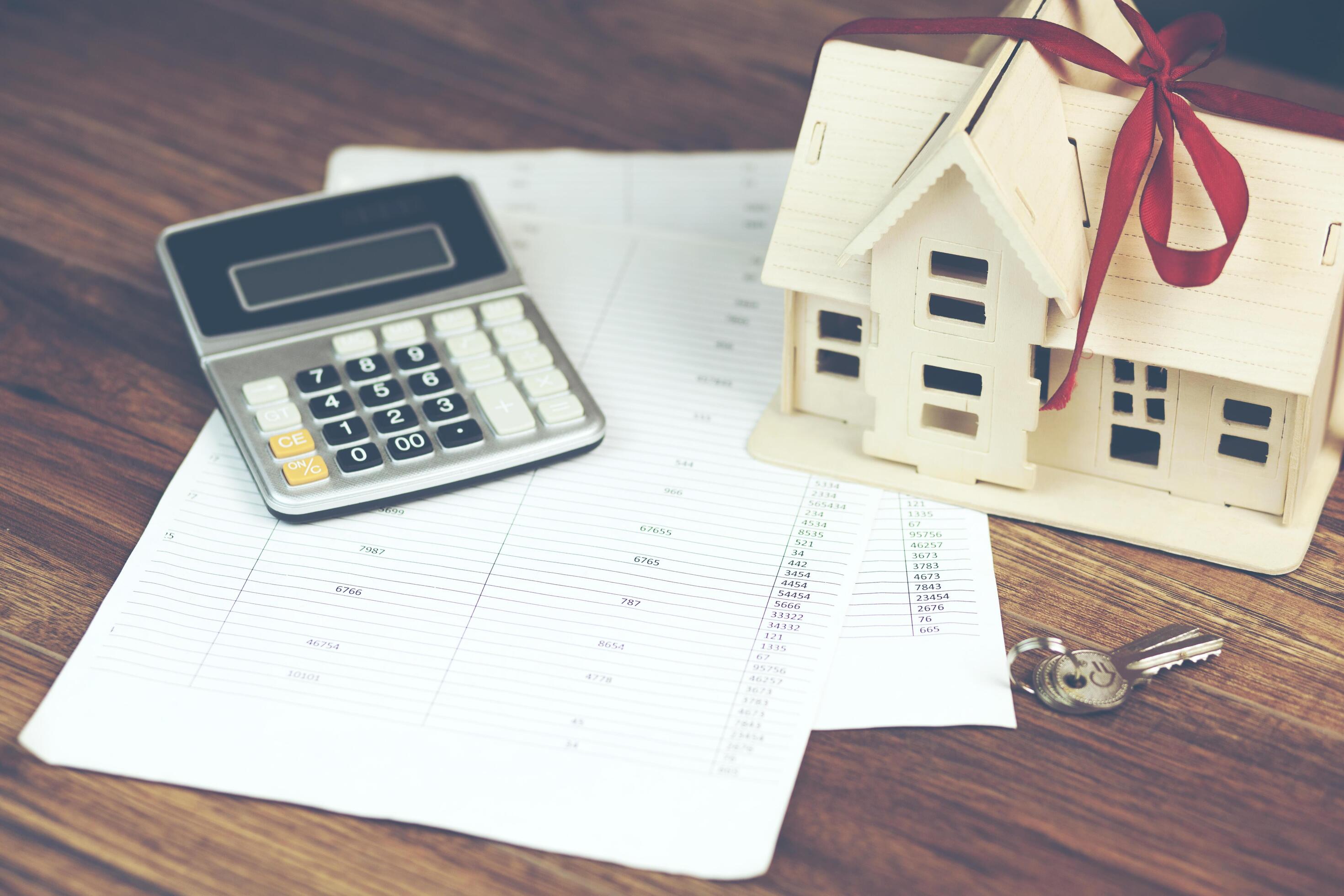 A calculator, paperwork, wood model of a house, and keys on a wooden table