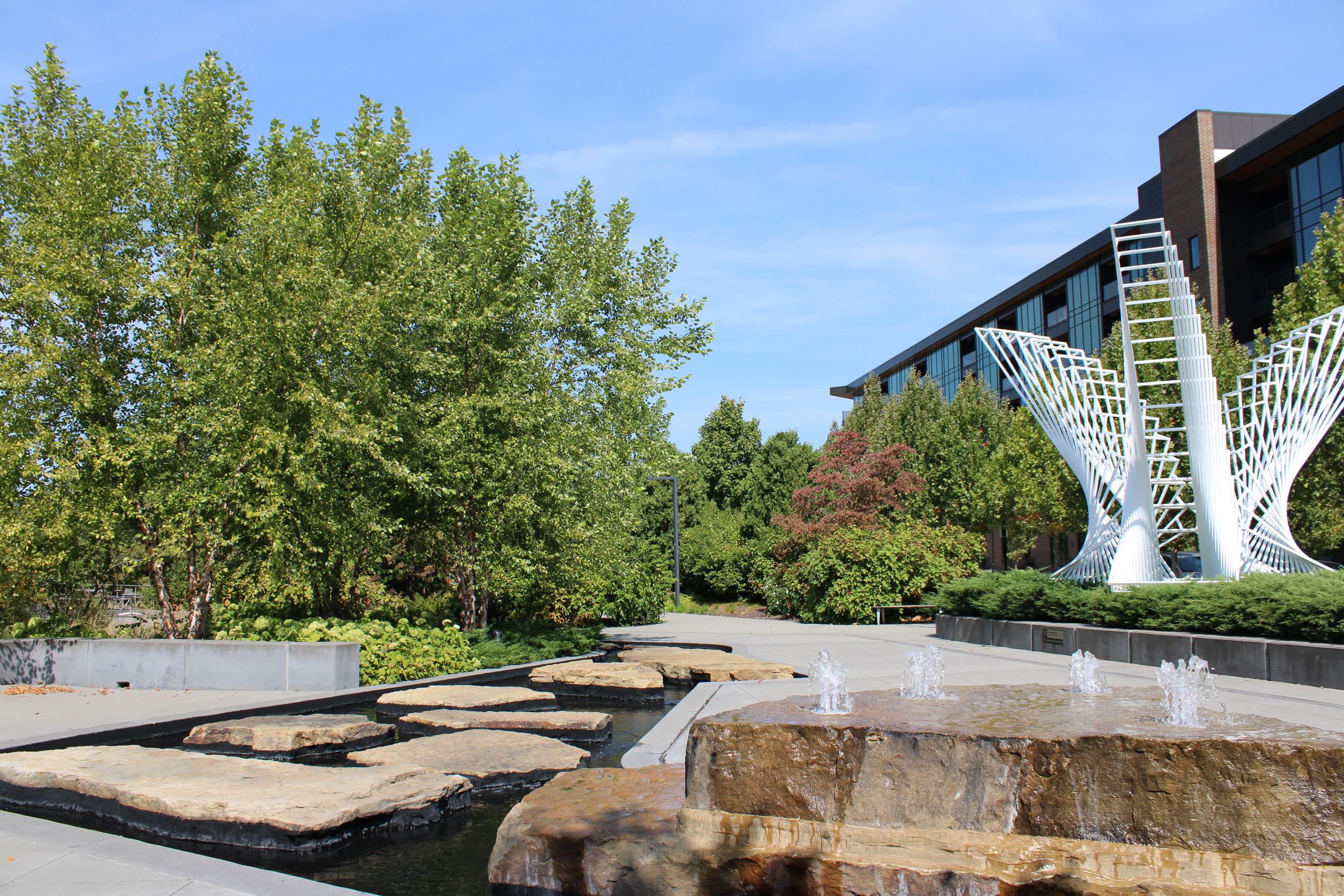 A landscaped area featuring a large white abstract sculpture, lush green trees, and a water feature with smooth stones and small fountains.