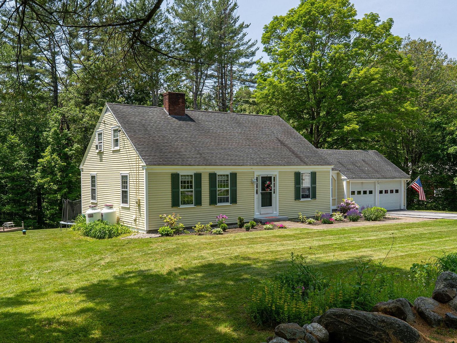 Front exterior of 26 Garrison Lane, a yellow Cape-style home in Hopkinton NH with green shutters, a stone wall, and perennial gardens