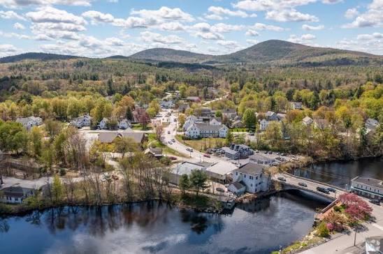 White New England house on a rocky waterfront with several smaller accessory outbuildings beside it and autumn foliage behind, a Goffstown NH ADU illustration