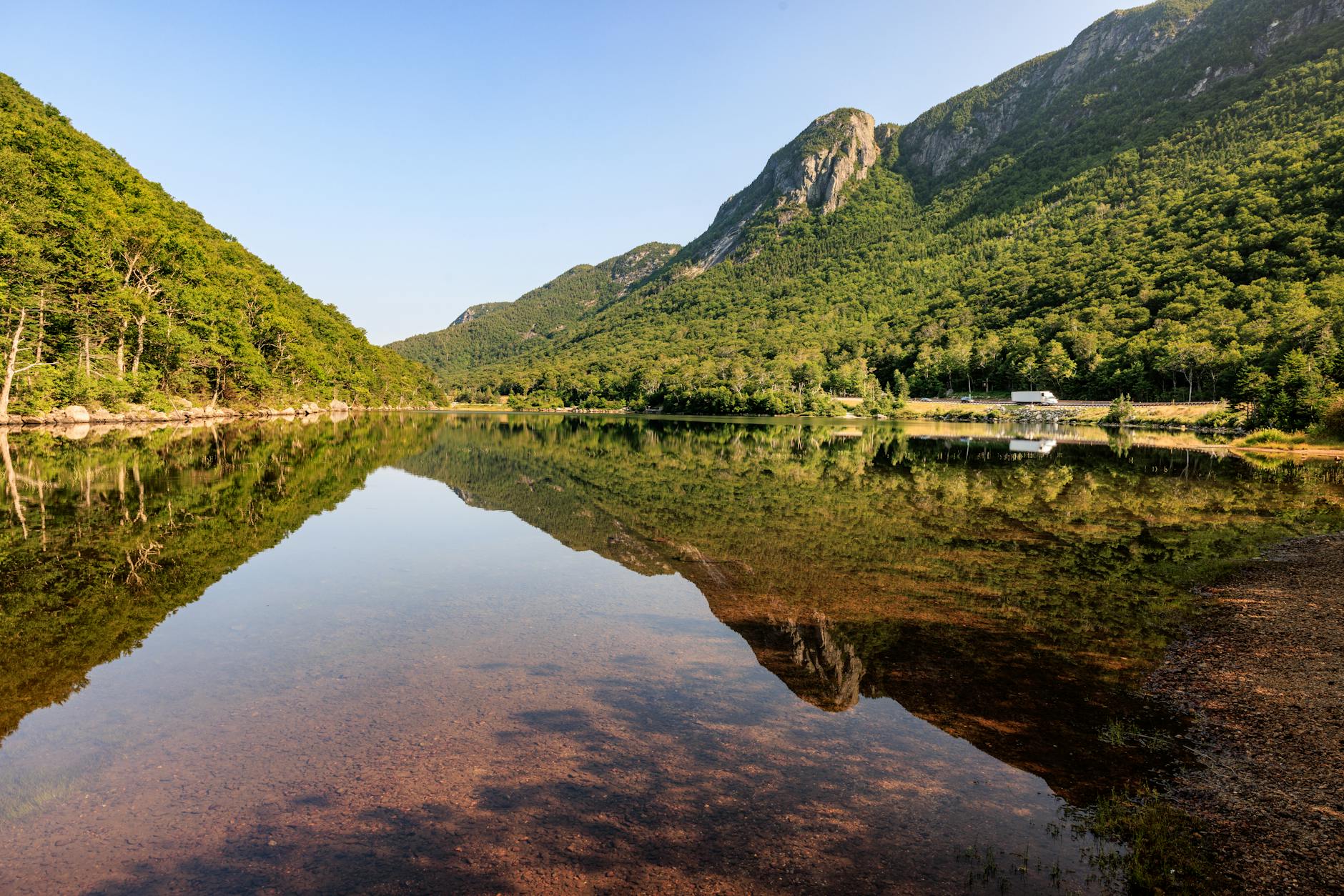 Glassy New Hampshire pond reflecting green forested hills on a clear summer morning — the outdoor lifestyle that draws home buyers to towns like Hollis NH