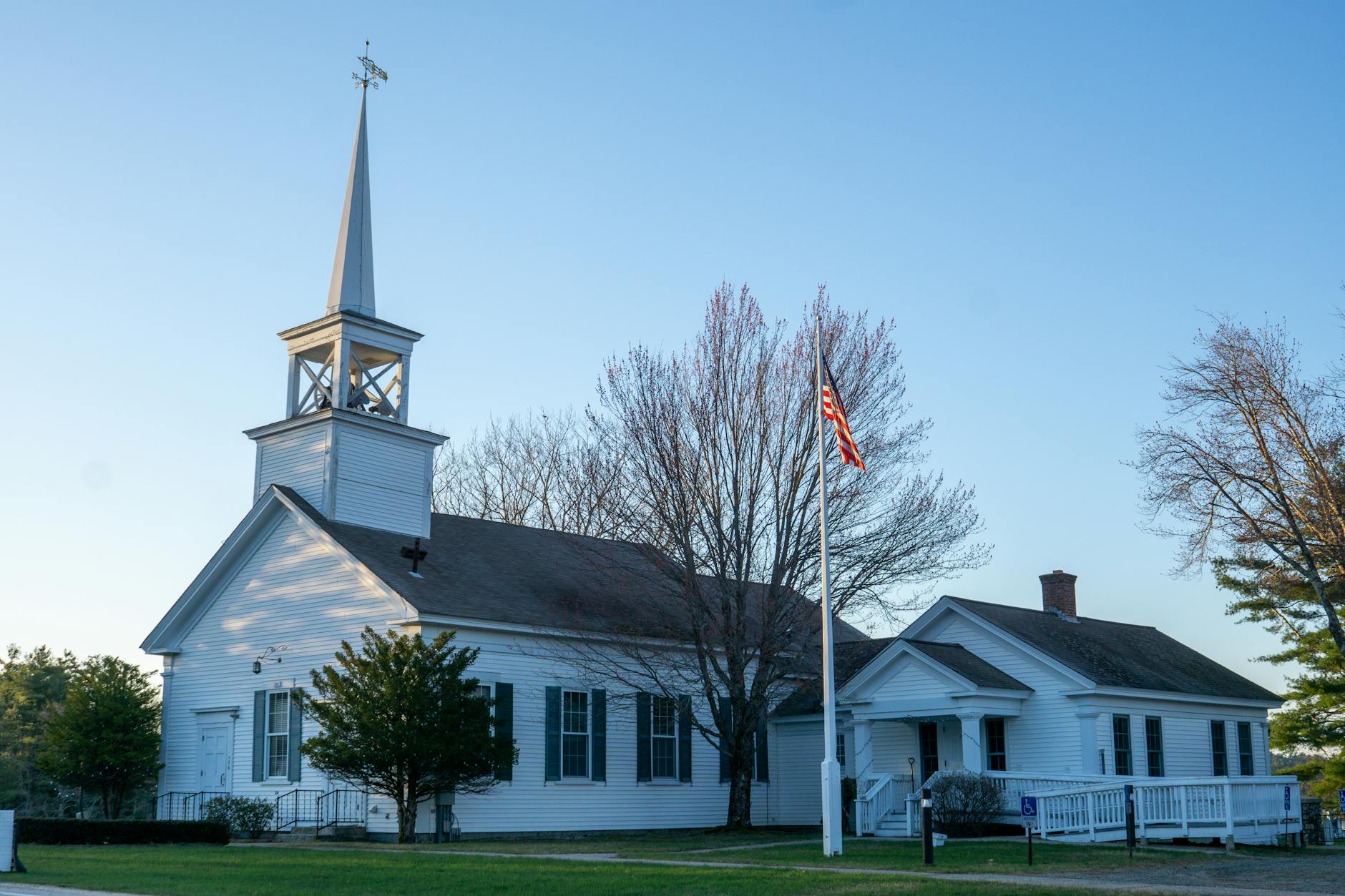 Historic white clapboard New England meetinghouse with tall steeple, weather vane, and American flag — symbol of the East Derry NH neighborhood and its First Parish Church