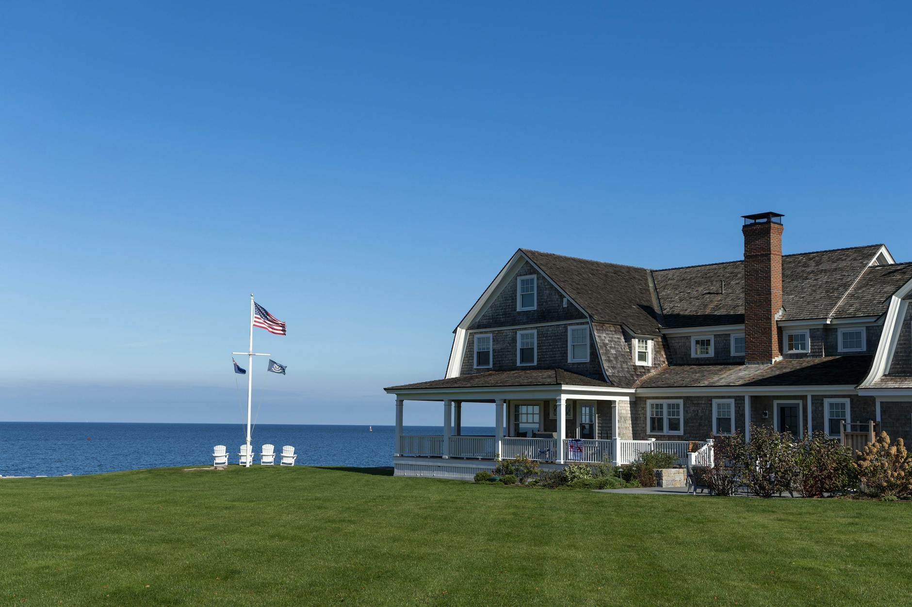 Waterfront New England shingle-style home with a wraparound porch and green lawn overlooking open water, representing lakefront homes for sale in New Hampshire
