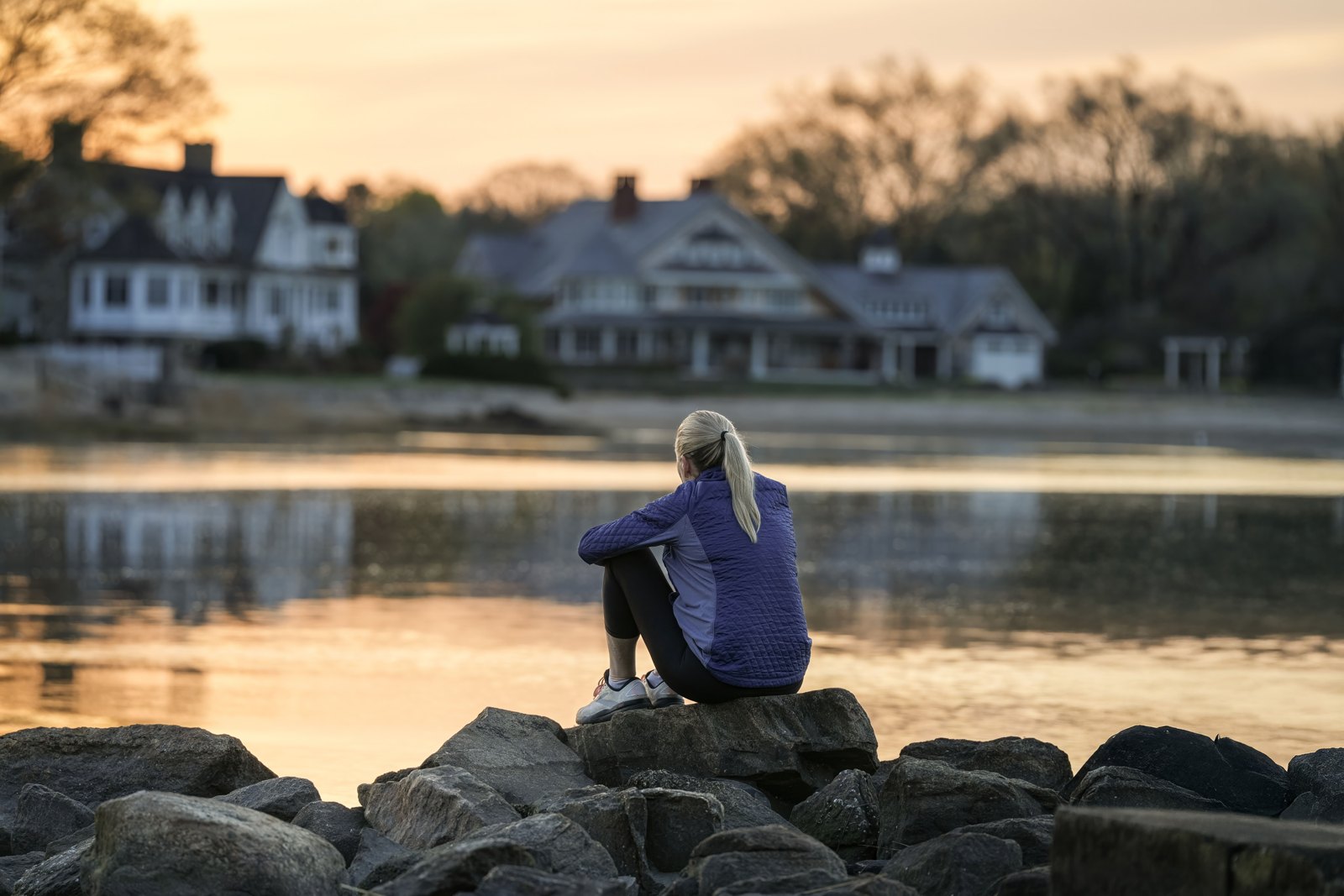 A woman sits on harbor rocks at dusk looking across calm water toward a row of large shingle-style waterfront homes on the New Hampshire seacoast