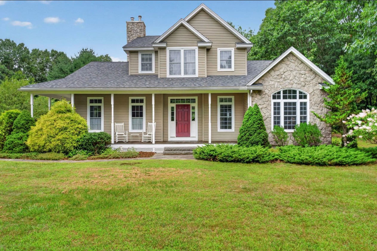 Front exterior of a tan clapboard home with stone accents and a covered farmers porch on a manicured lawn