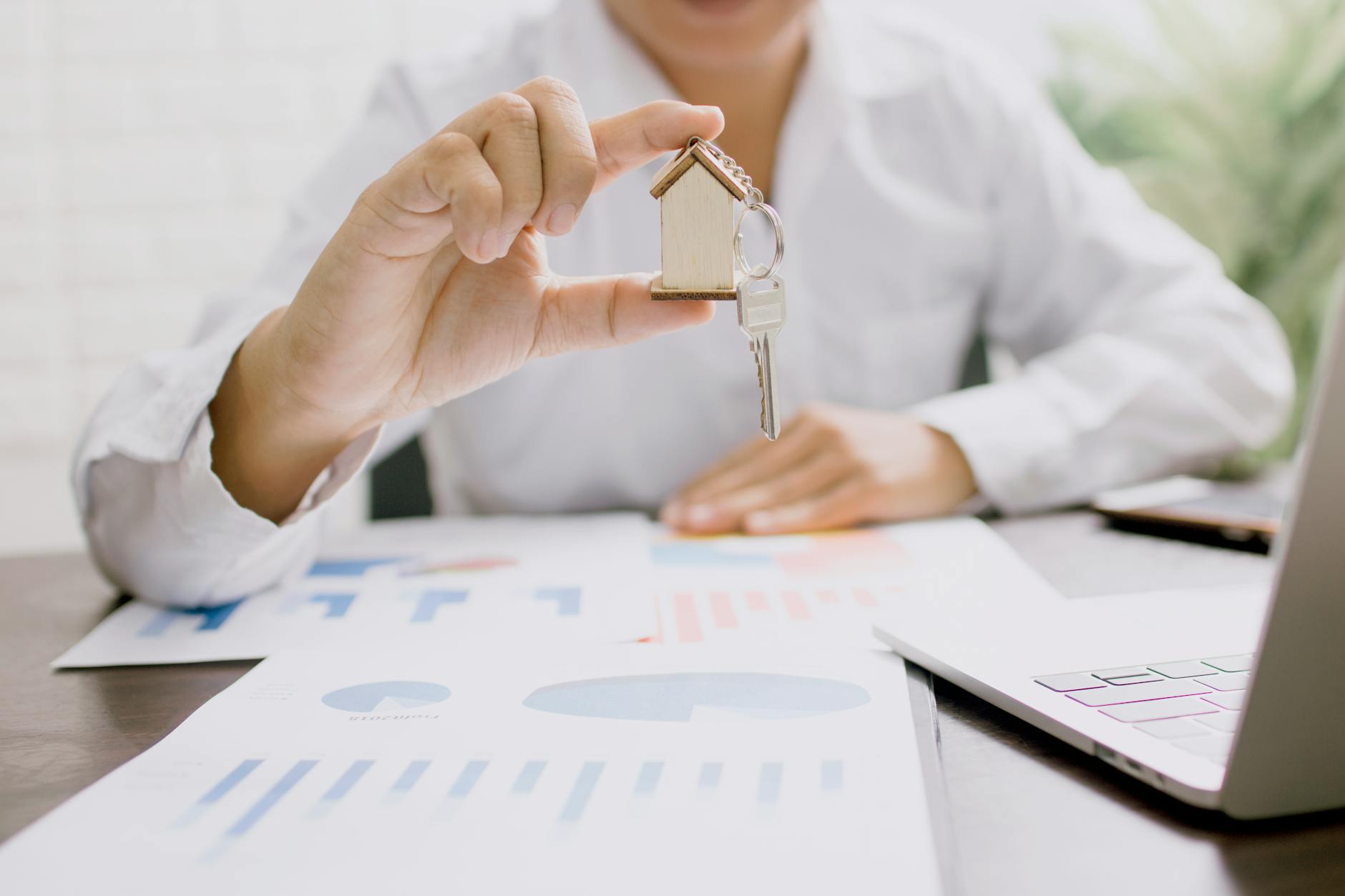 Person holding a house-shaped keychain and key over market charts and a laptop at a desk — planning a move to Concord NH
