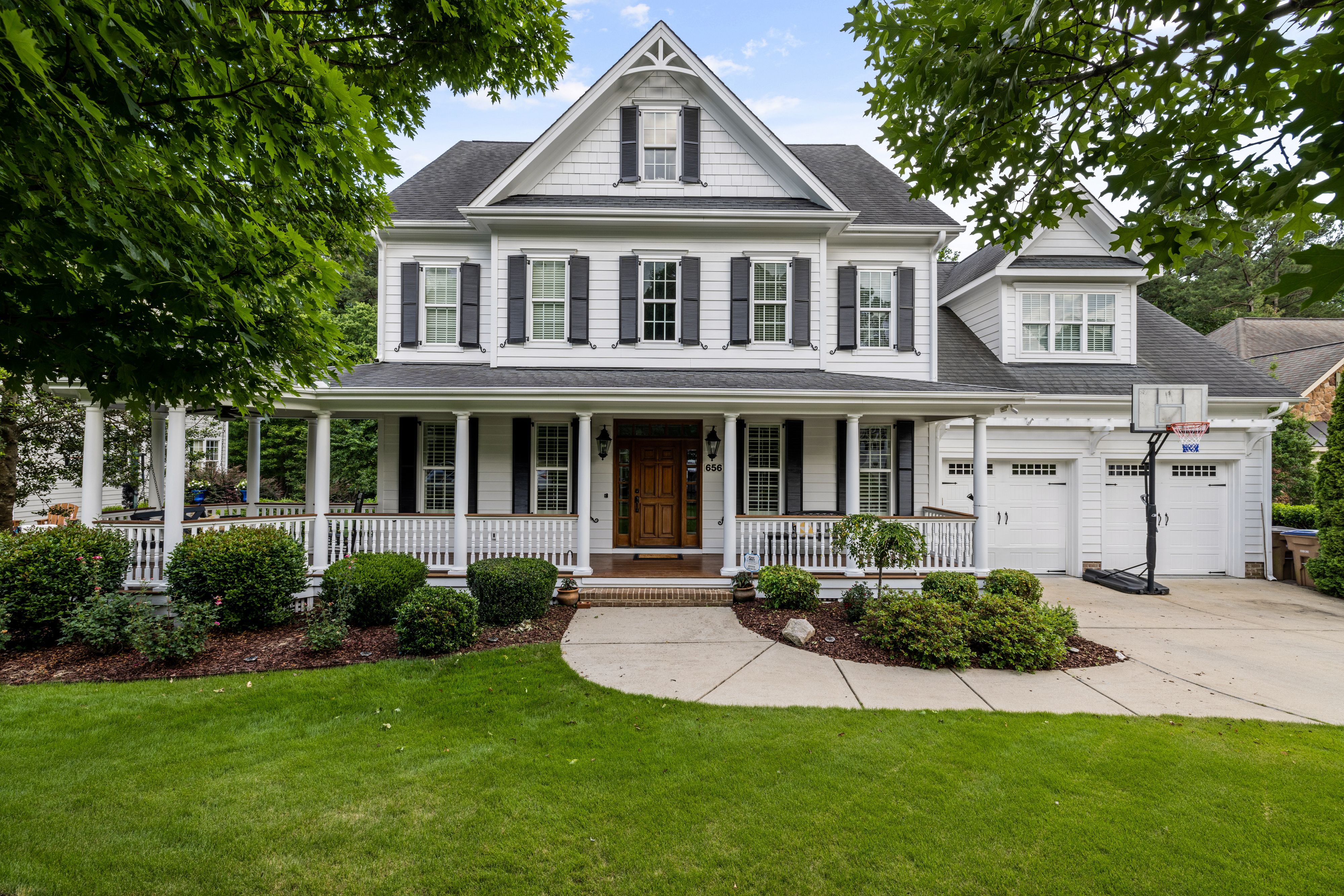 White farmhouse with a wraparound porch and attached garage on a landscaped summer lawn, a family home near Concord NH schools