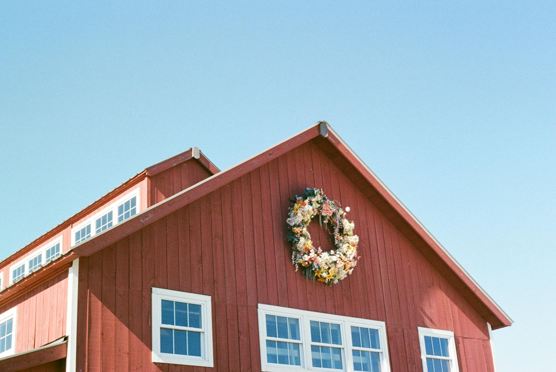 Red barn-style New England building with white-trimmed windows and a floral wreath under a clear summer sky