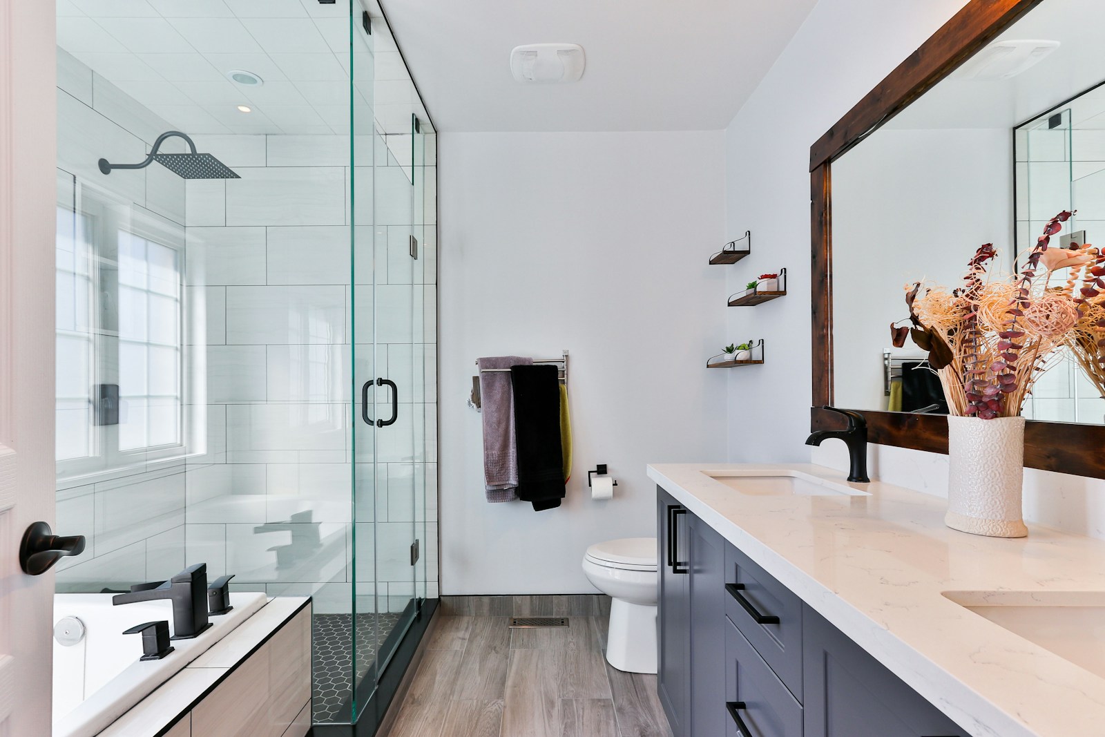 Bright renovated bathroom with frameless glass shower, navy double vanity with quartz counters, matte black fixtures, and wood-look floors